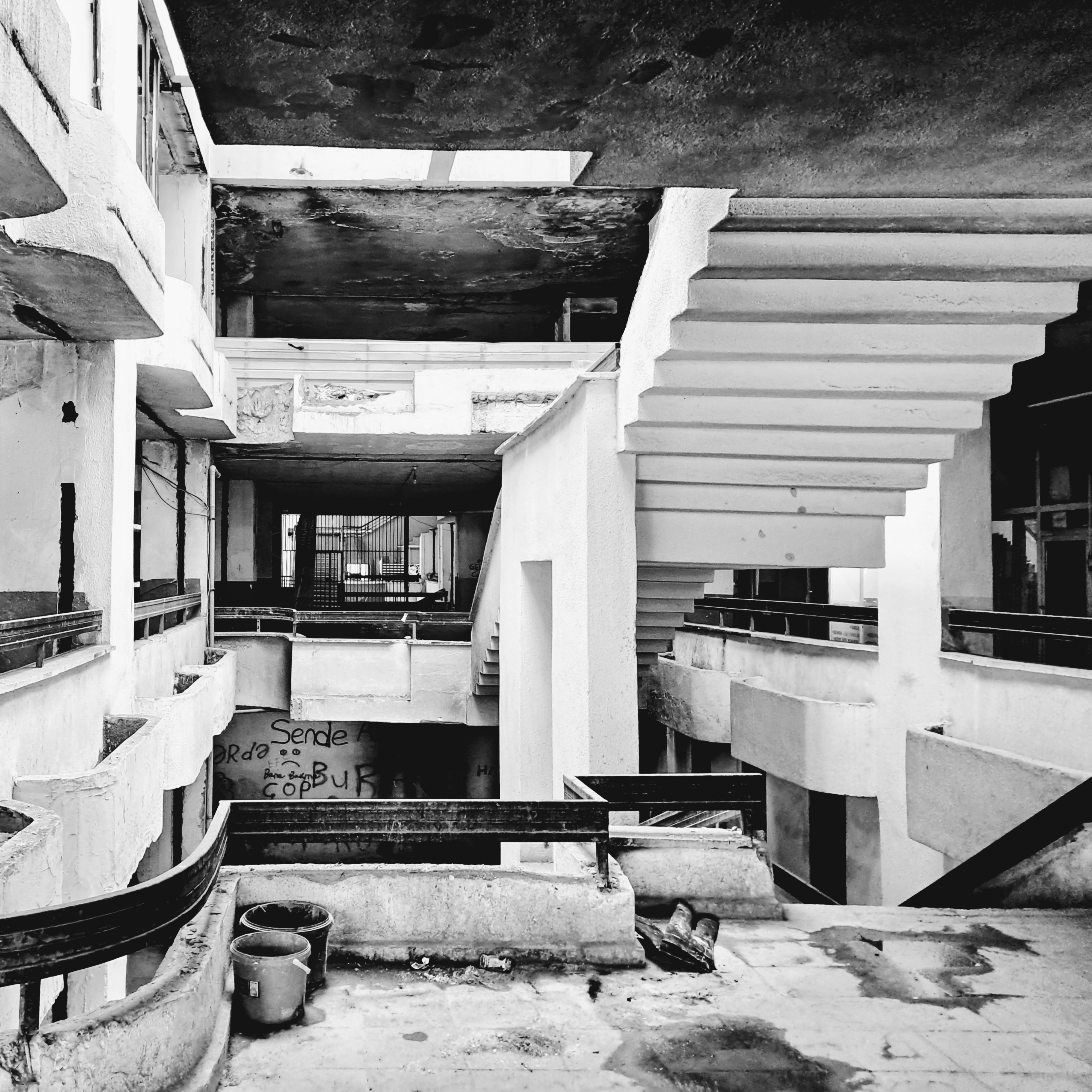 A view looking through the multi-level interior of a dilapidated brutalist concrete building. Prominent concrete staircases with angular undersides lead upward alongside open balconies with dark railings and weathered surfaces. Debris, water stains, and two discarded buckets are on the tiled floor in the foreground, while damaged ceilings expose faded concrete above. Hand-drawn graffiti on a wall across the central opening reads "Your" above a sad face symbol, accompanied by text that appears to read "There" and "trash IS HERE". Natural light enters into the structure from upper openings and an iron security gate is visible deep in the background corridor.