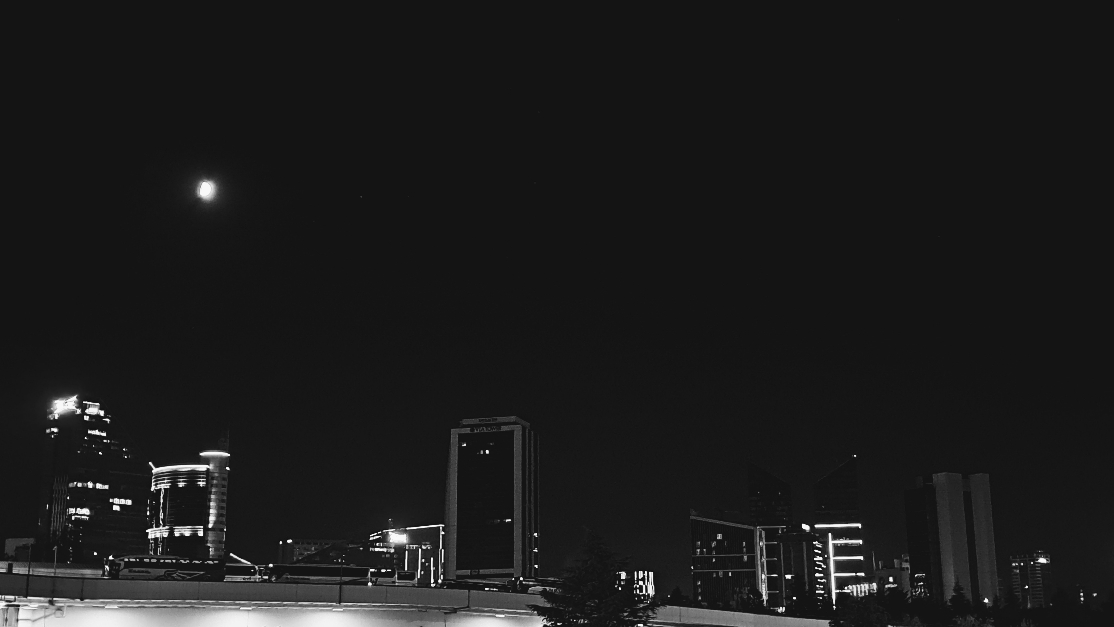 A black and white nighttime cityscape under the dark sky with the moon shining above. Below, the silhouettes of modern high rise buildings and skyscrapers stand out with illuminated windows and glowing architectural details. In the immediate foreground, a brightly lit elevated ramp stretches across the view, with a few buses strolling through.