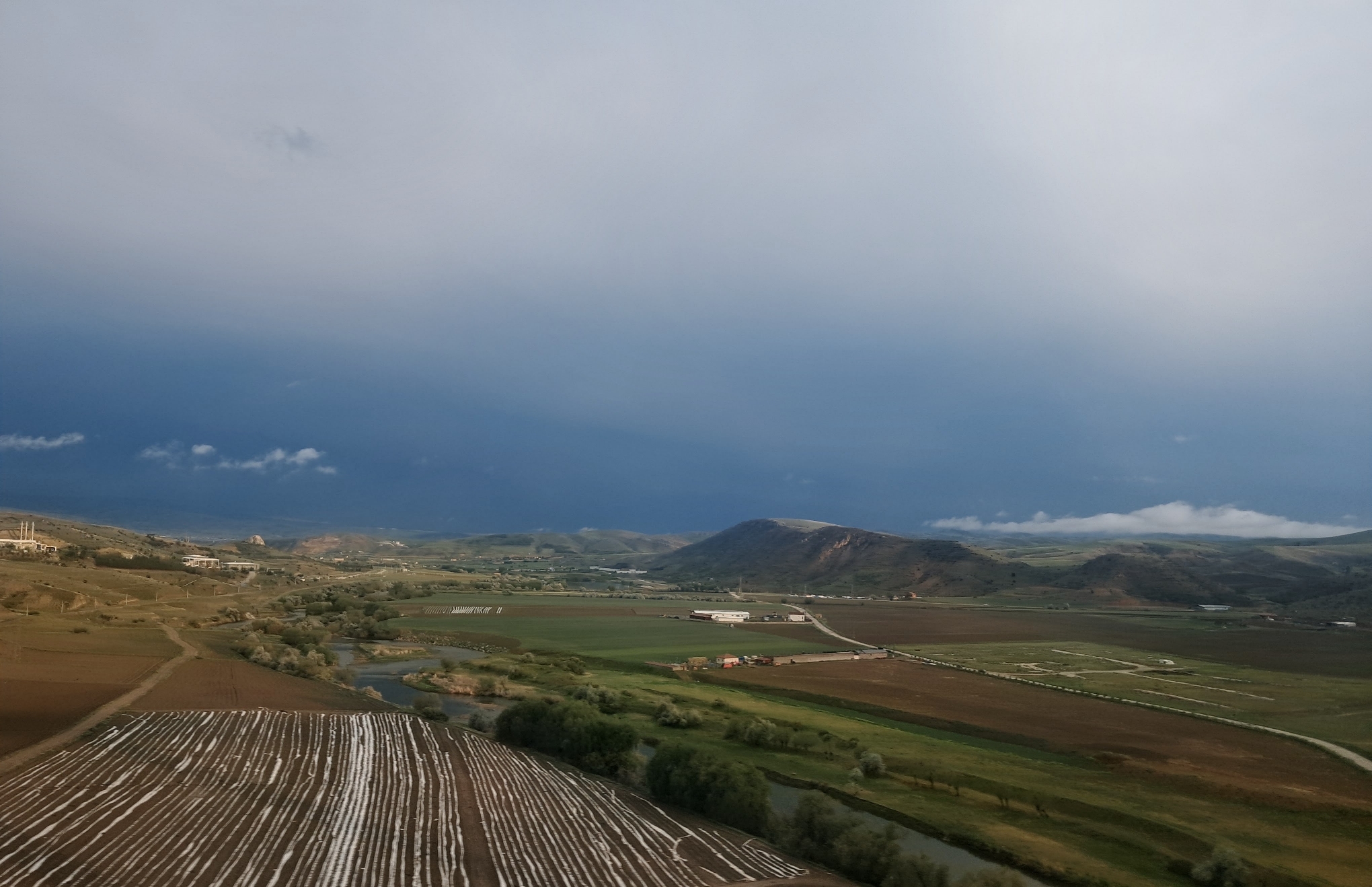 Aerial view of a rural valley under an overcast sky filled with storm clouds. In the foreground, a plowed agricultural field features parallel white mulch coverings for crops. A winding river lined with trees cuts through the valley floor, separating the fields of green and brown. In the distance, rolling hills rise beneath low clouds, with a cluster of industrial buildings in the landscape.