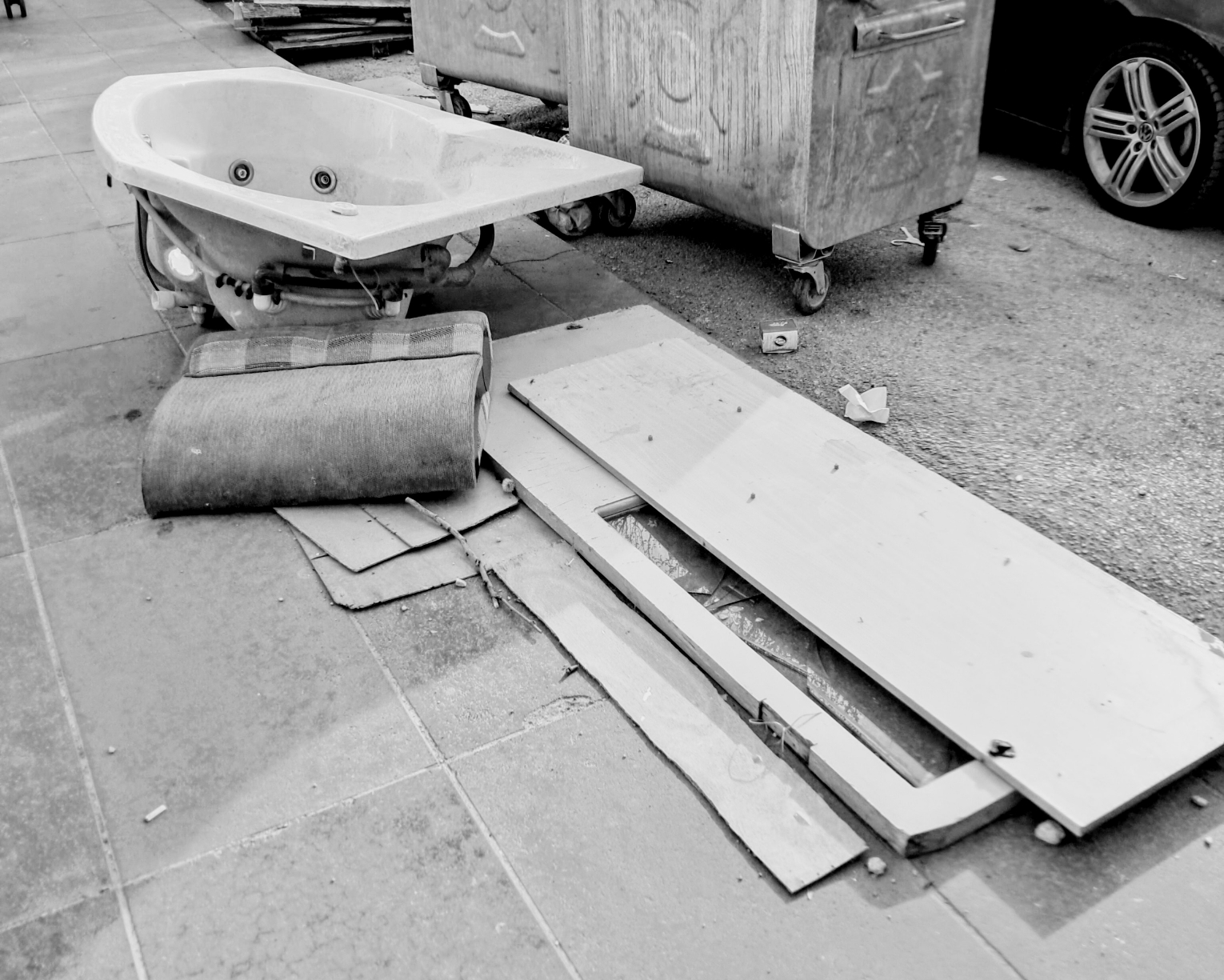 Discarded household and renovation debris left on a sidewalk next to large metal dumpster bins and the front wheel of a parked car. A white jacuzzi-style bathtub with visible underside plumbing rests on the pavement beside a piece of a sofa. In the foreground, flat wooden boards and a disassembled door frame lie across pieces of cardboard and scattered street litter.