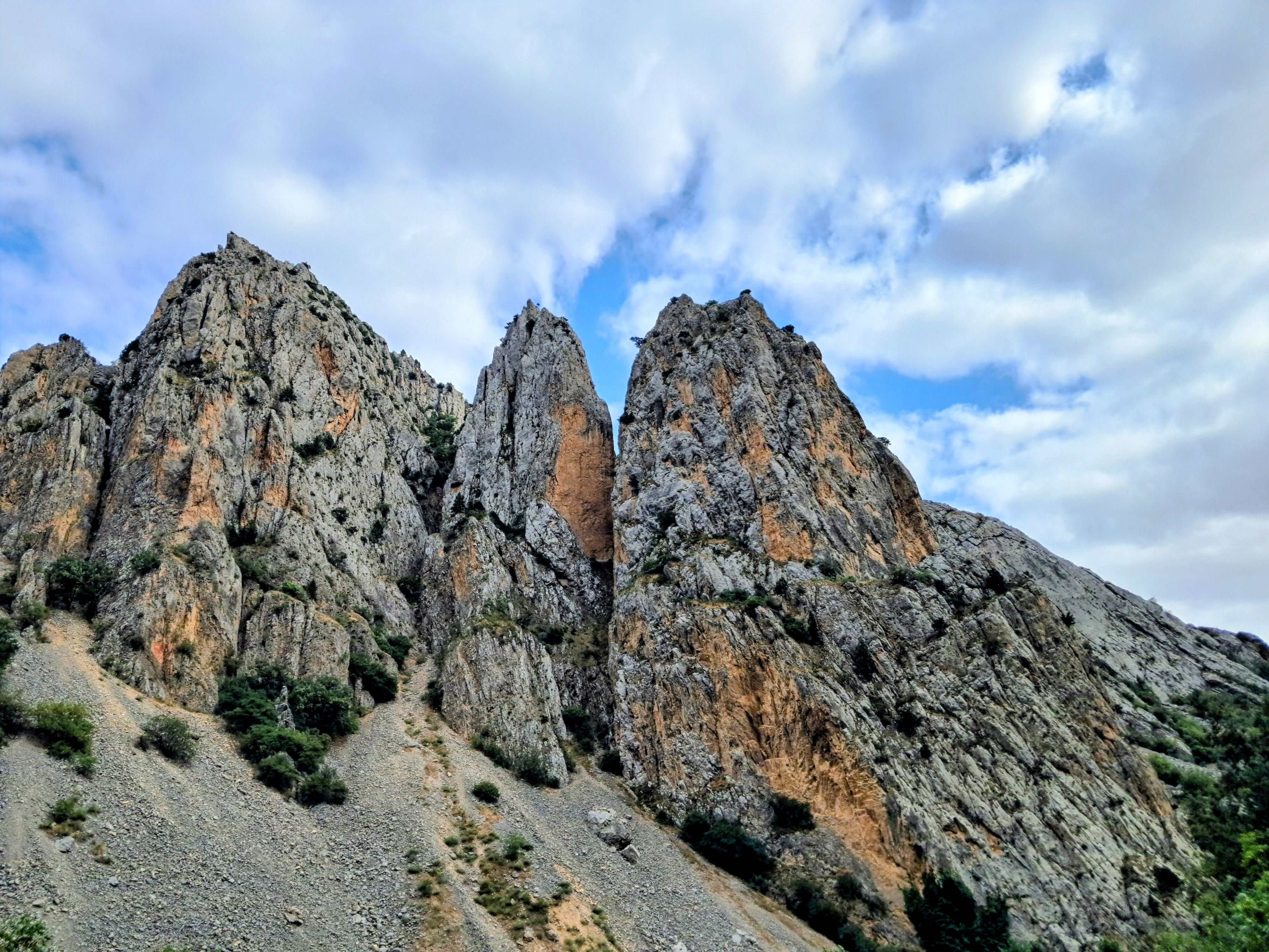 Low-angle view of rugged limestone rock formations rising steeply into a bluesky with heavy clouds. The jagged cliffs feature a mix of gray stone and warm orange patches, with sparse green shrubs appearing among the crevices and loose scree covering the lower slopes.