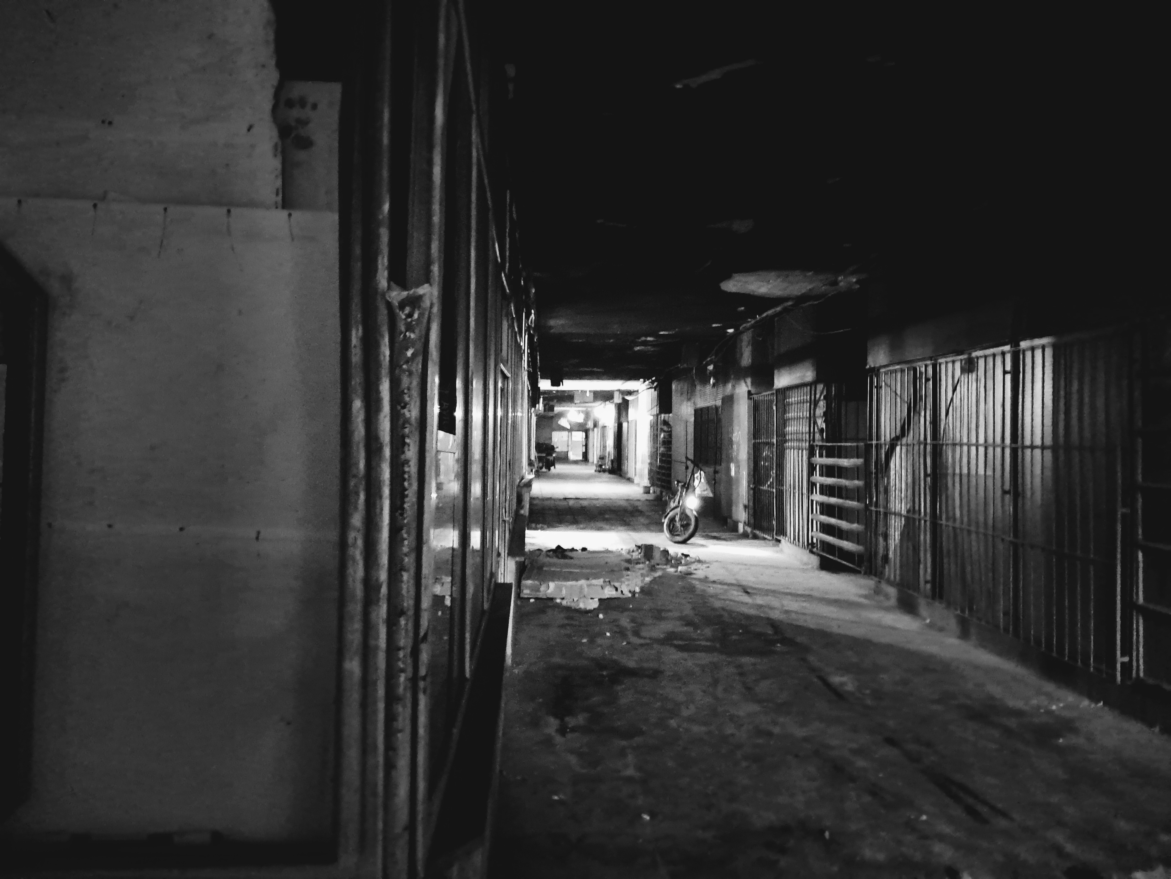 A rundown enclosed corridor lined with weathered walls and closed metal security gates along the way. In the middle distance, an electric scooter sits parked, illuminated under a patch of light next to scattered debris on the floor. The ceiling is also damaged and peeling, leading the eye down the gloomy hallway toward a brightly lit opening in the far background.