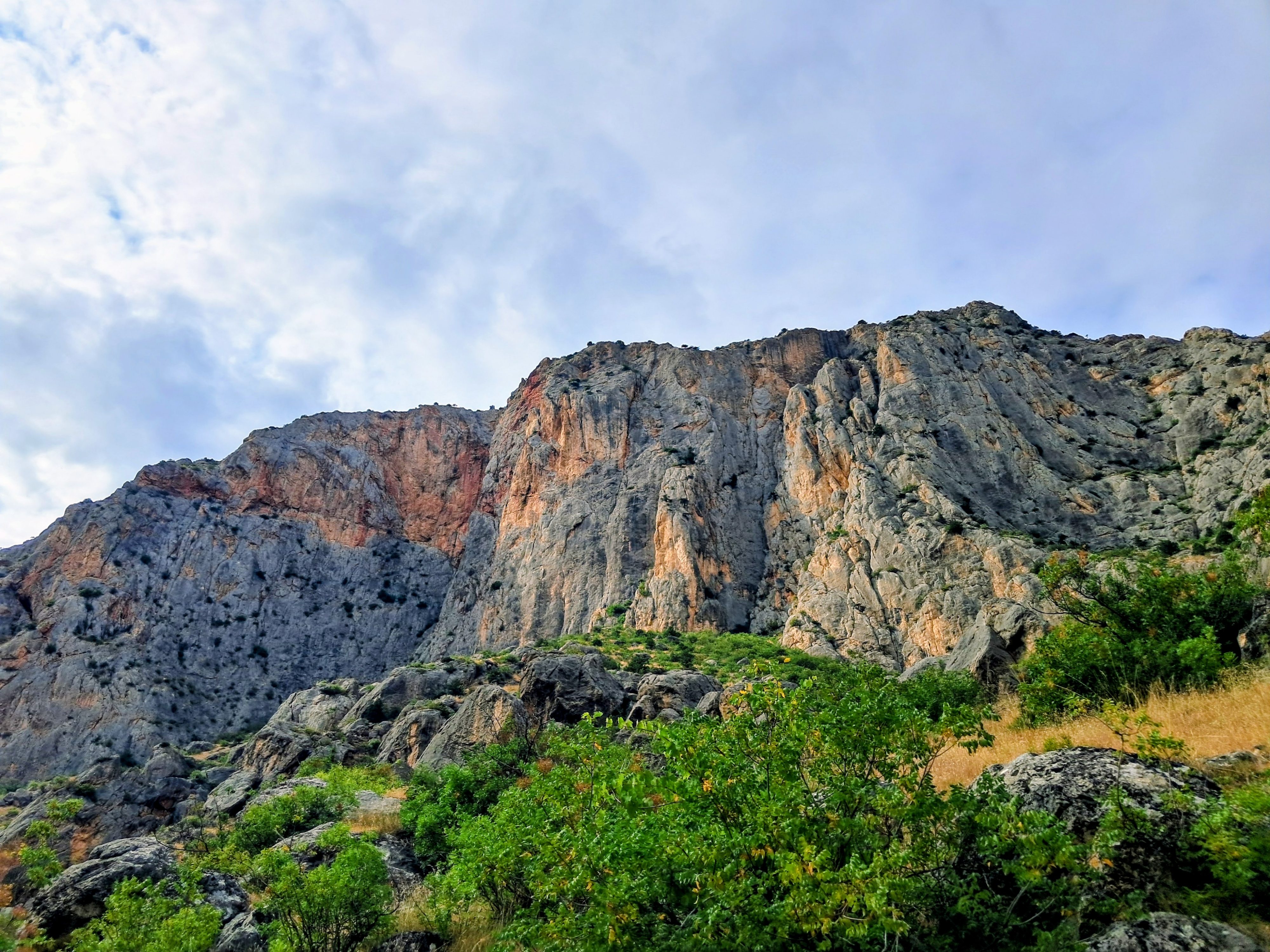 Large steep rocky mountain with exposed vertical cliff faces, rugged gray and tan stone formations, green vegetation and dry grass along base and lower slopes under partly cloudy blue sky.