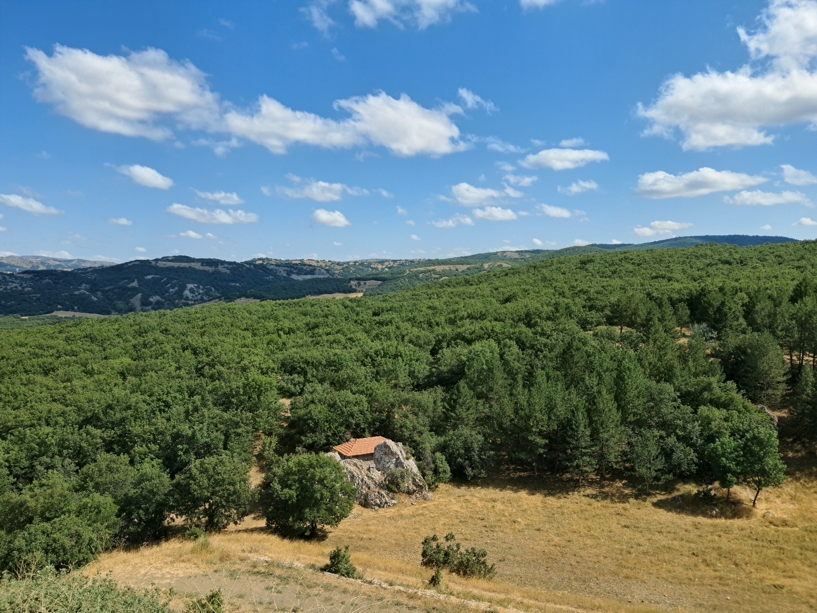 A view of expansive green forested hills with small house featuring red roof built into rocky outcrop in foreground clearing, under bright blue sky with scattered white clouds.
