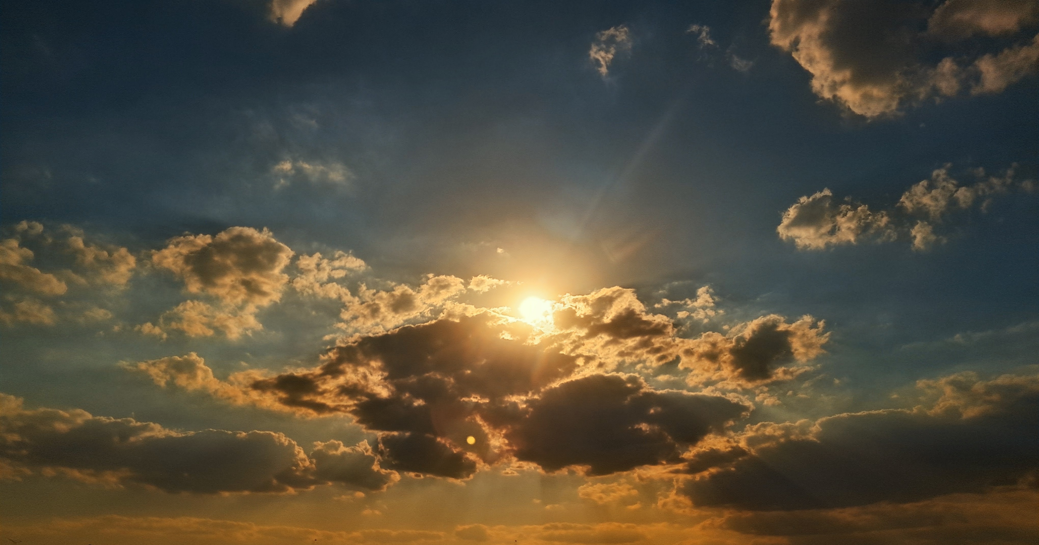 Bright sunlight bursts from behind a dark central cloud formation, casting sunbeams upward and downward across a wide sky. The upper sky shifts to deep blue, while scattered puffy clouds near the horizon and surrounding the sun are illuminated with warm orange, golden, and reddish highlights.
