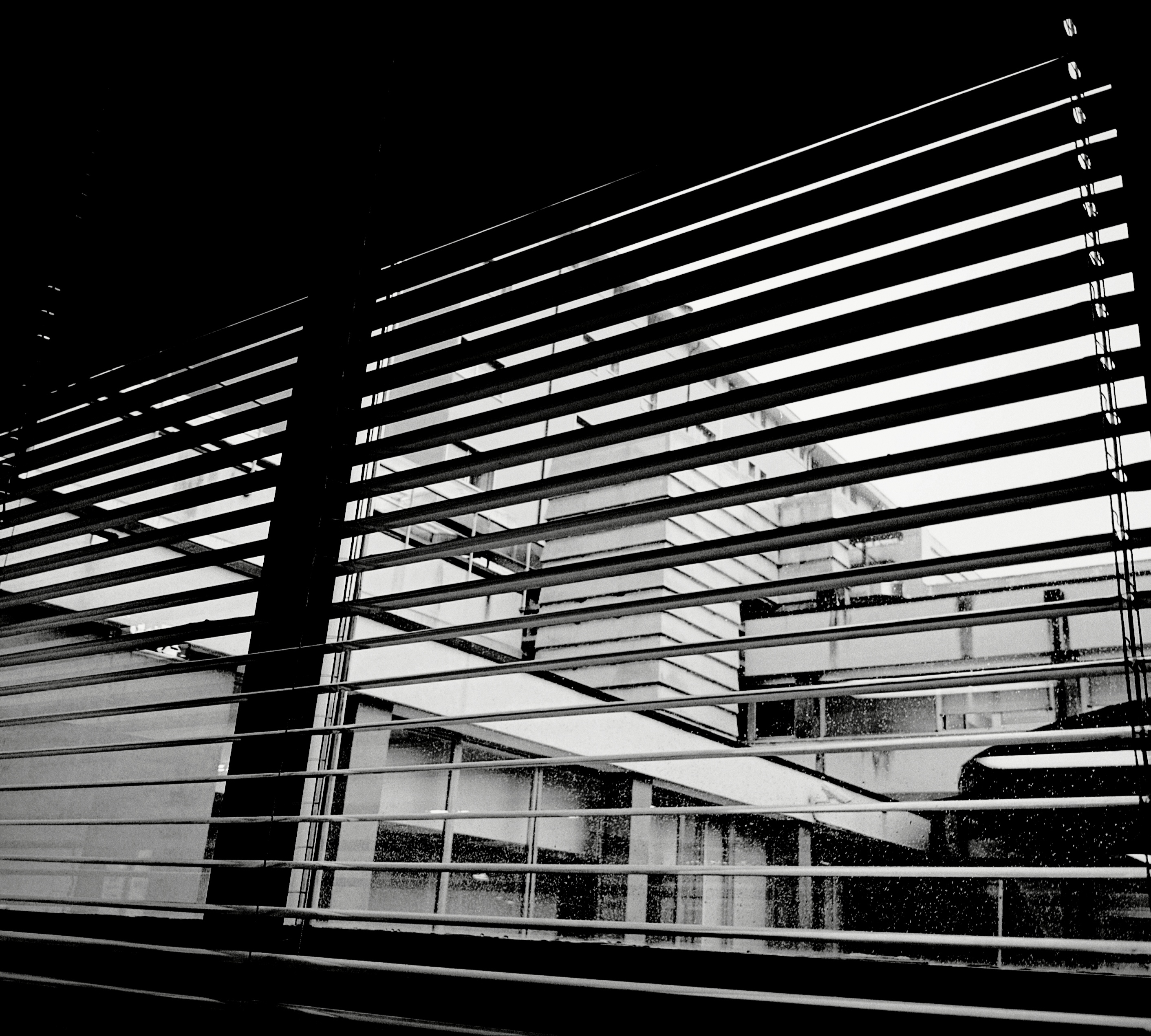 Una finestra, parzialmente oscurata da una veneziana, lascia intravvedere un edificio all'esterno. Foto in bianco e nero.