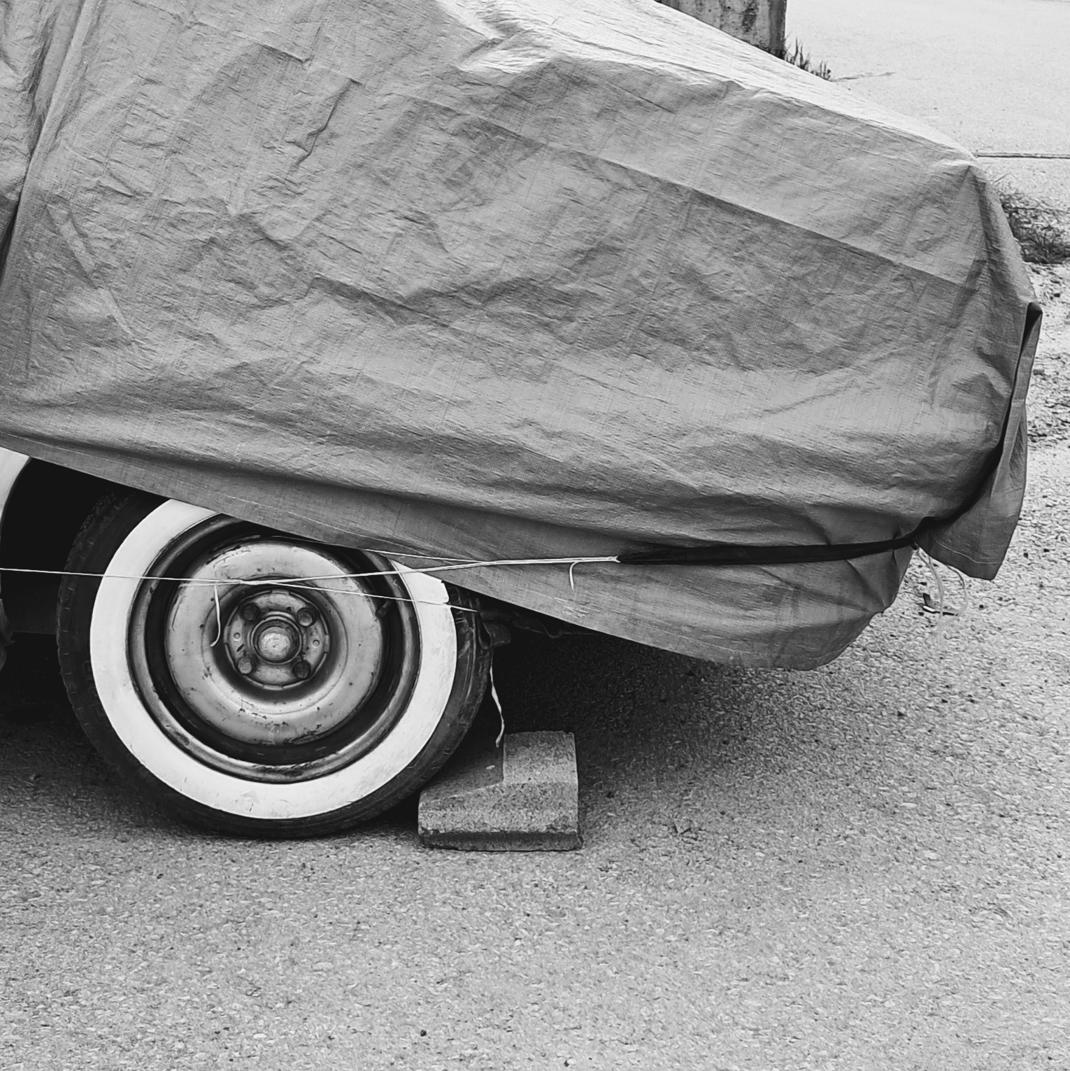 The rear quarter of a vehicle covered by a rumpled fabric tarp secured by straps. The rear wheel is visible, featuring a tire with a prominent wide whitewall and a metal rim, with a concrete block placed behind it. A small portion of a building, paved road, and some natural grass are in the background.