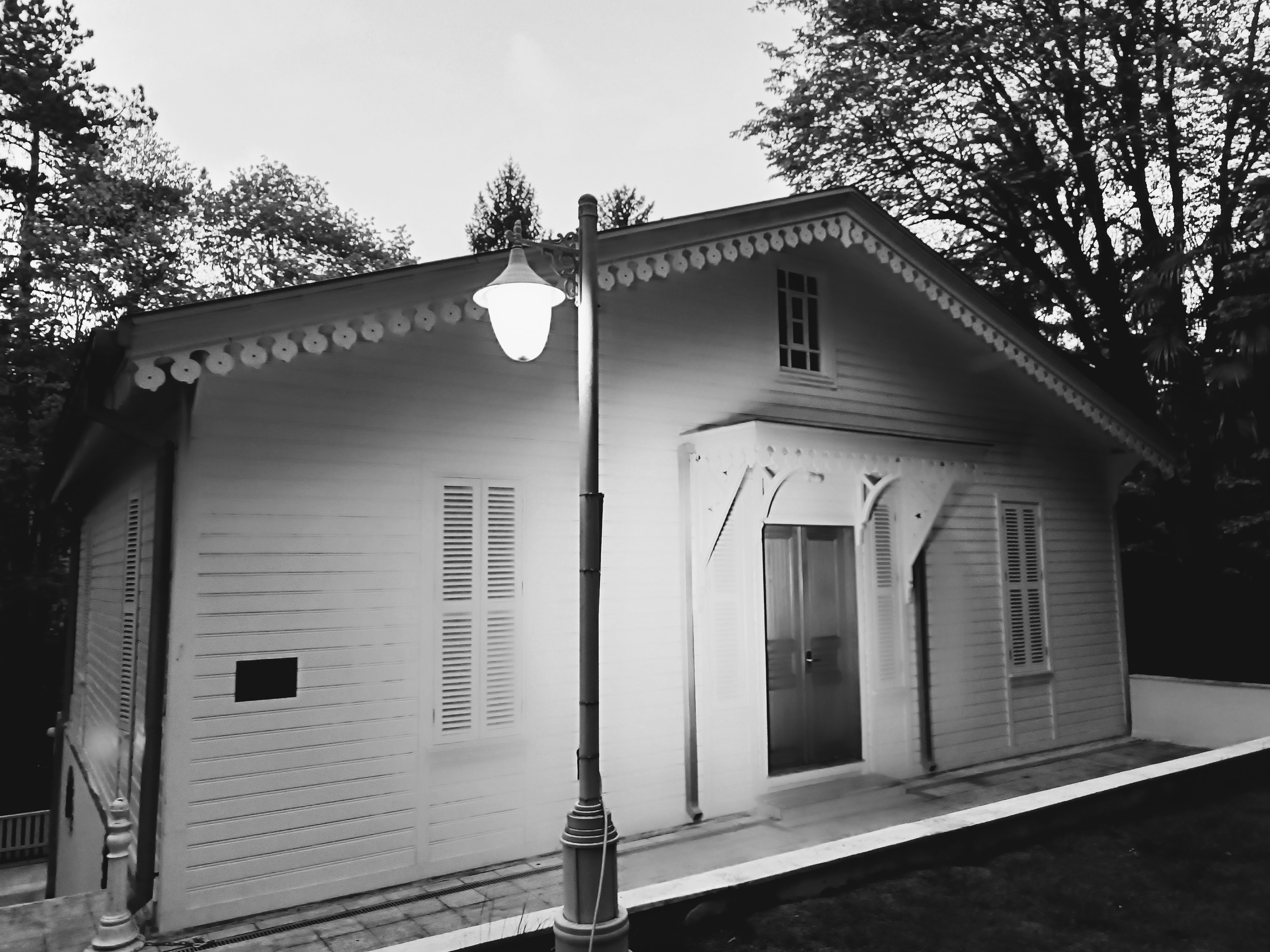 A single-story wooden cottage with decorative gingerbread trim under its gabled roof and an ornate canopy over the central glazed spruce door. Shuttered windows are placed along the entrance, and an illuminated vintage street lamp stands along the stone path in the foreground. A small rectangular plaque is mounted to the left exterior wall with faded text. Dense trees rise in the background behind the building.