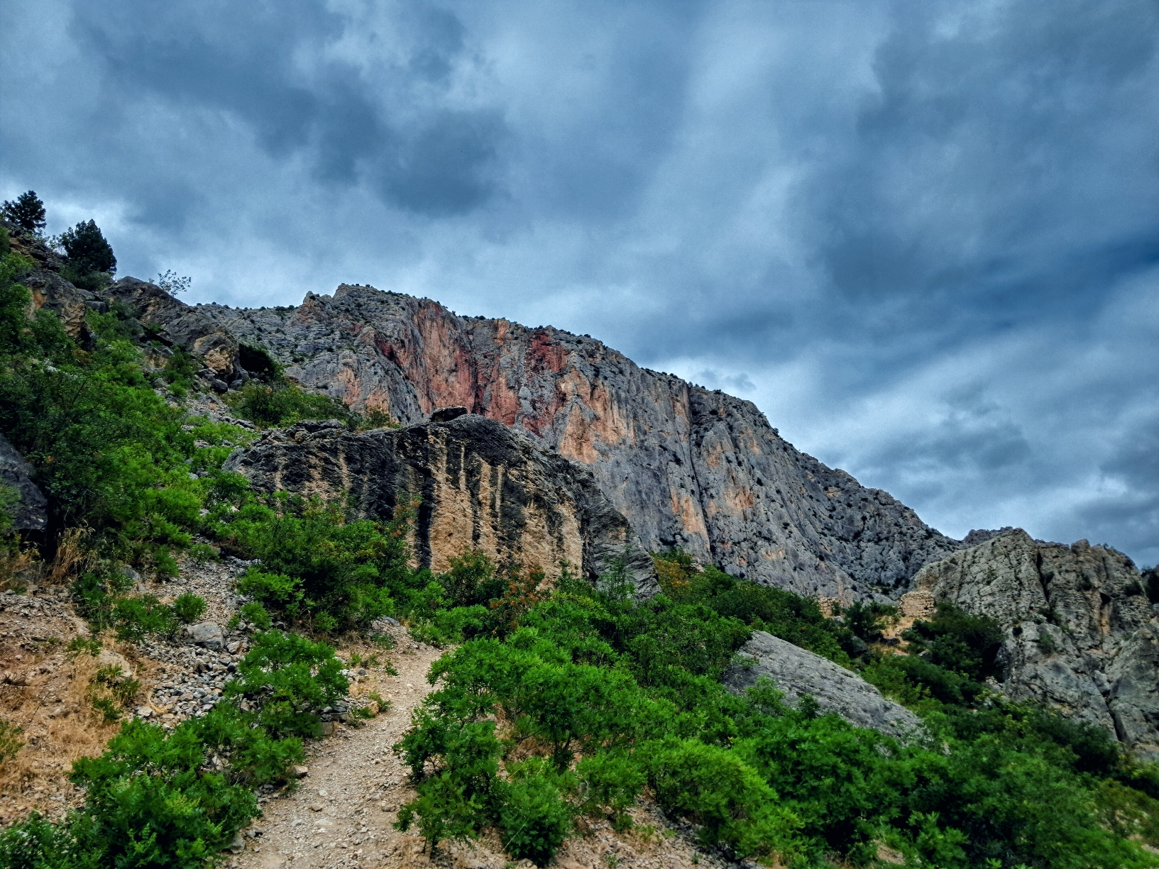 A narrow dirt hiking path goes upward through green shrubs and scattered rocky terrain toward steep craggy cliffs. The rock face rises prominently across the background, featuring shades of gray and reddish-brown stone. Above the mountains, dark storm clouds fill the blue and gray sky.