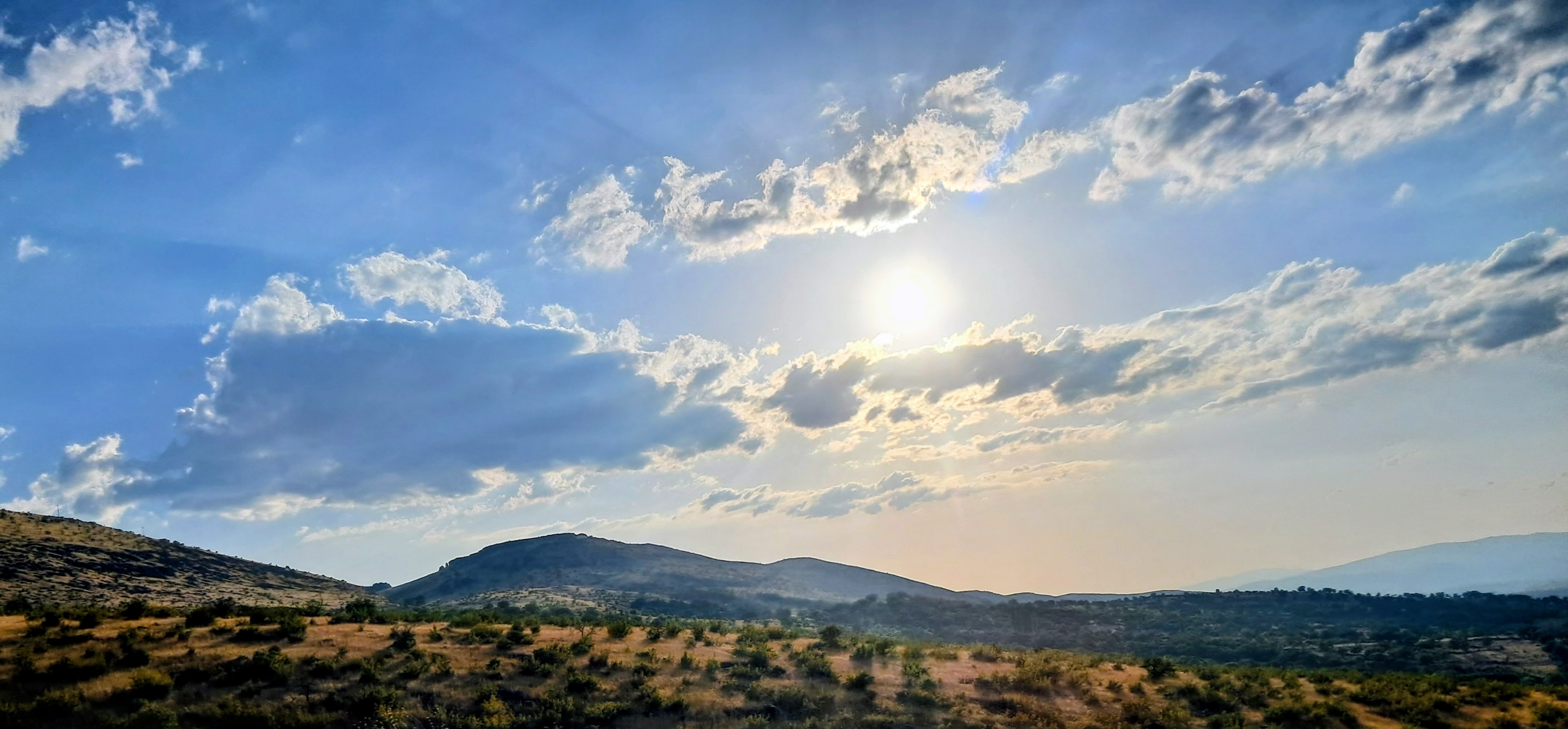 A view of rolling hills covered in dry yellowish grass and scattered low shrubs across the foreground towards a rounded mountain in the center background under the blue sky with scattered clouds Distant hazy ridges appear along the horizon under the warm afternoon light caused by the setting sun.