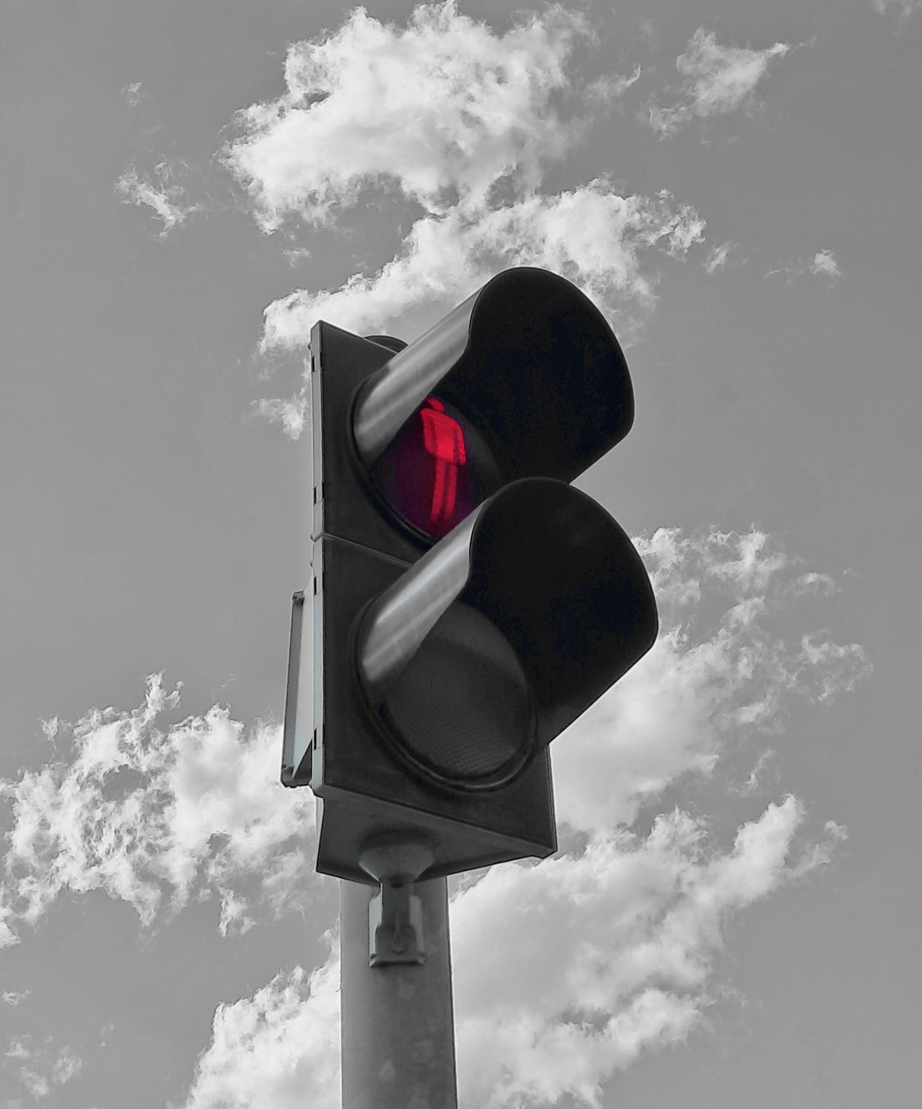 A pedestrian traffic light viewed from a low angle against a cloudy sky. The top light is illuminated in bright red, displaying a standing figure to indicate pedestrians should not cross. The bottom light is unlit, and the entire scene is in black and white except for the red light, creating a selective color effect.