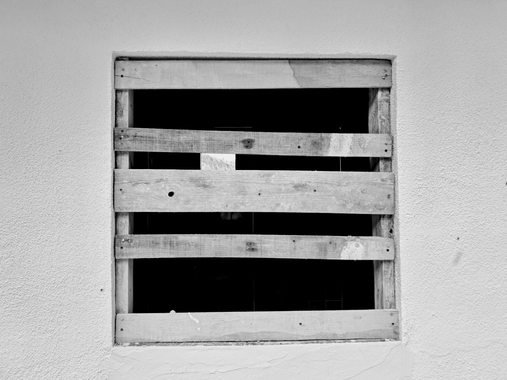 A square window opening set into a textured white exterior wall, boarded up with five weathered horizontal wooden planks nailed across the frame. The interior behind the gaps is mostly pitch black, with a small window opening visible in the upper center, reflecting light.