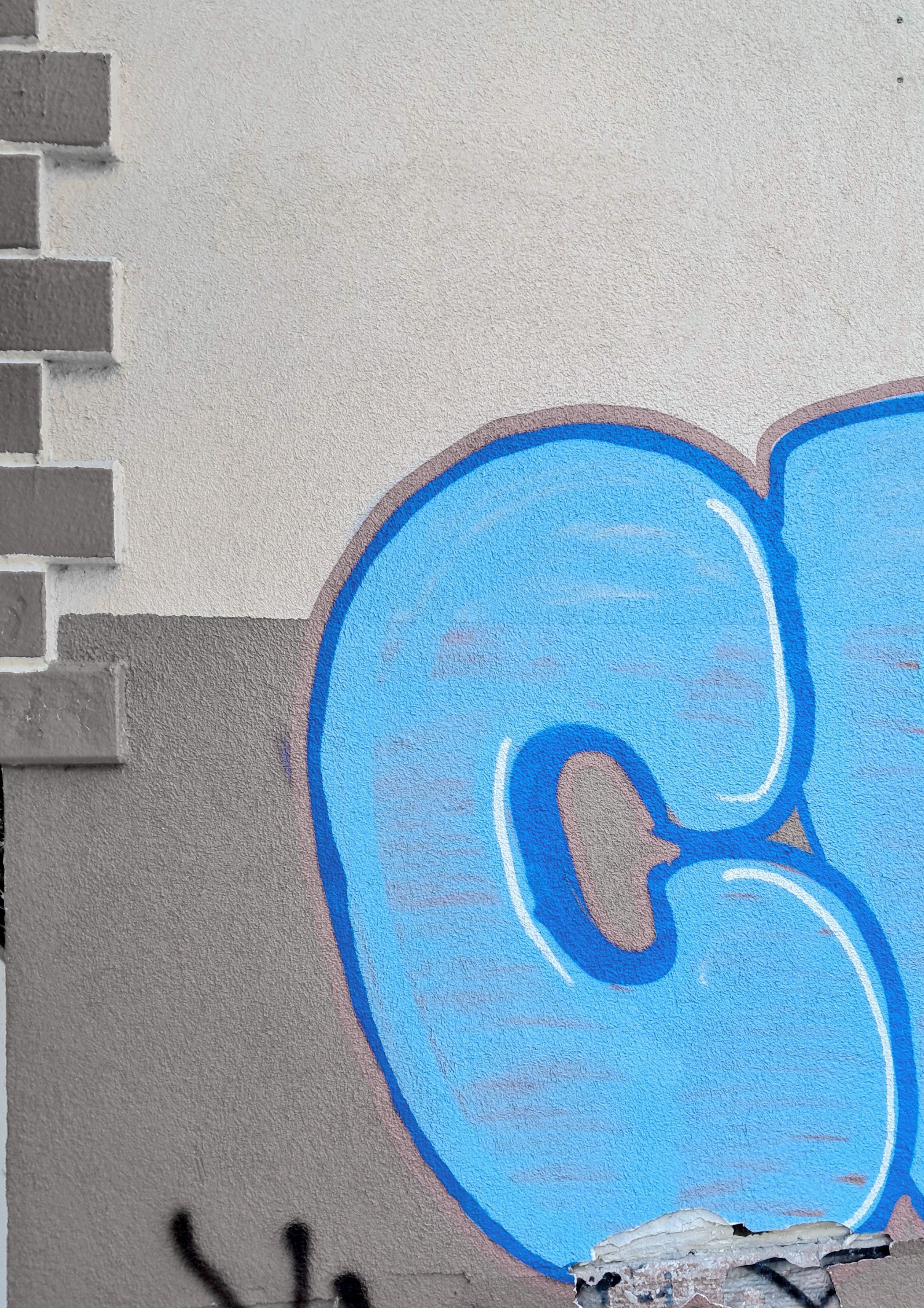 A textured outdoor wall features a large graffiti piece painted with bright blue bubble letters outline-highlighted in dark blue and thin white lines. The visible part of the lettering shows what appears to be a large letter C. The wall background transitions from a lighter off-white texture on top to a beige tone on the bottom, bordered by decorative vertical brickwork pattern on the left edge.