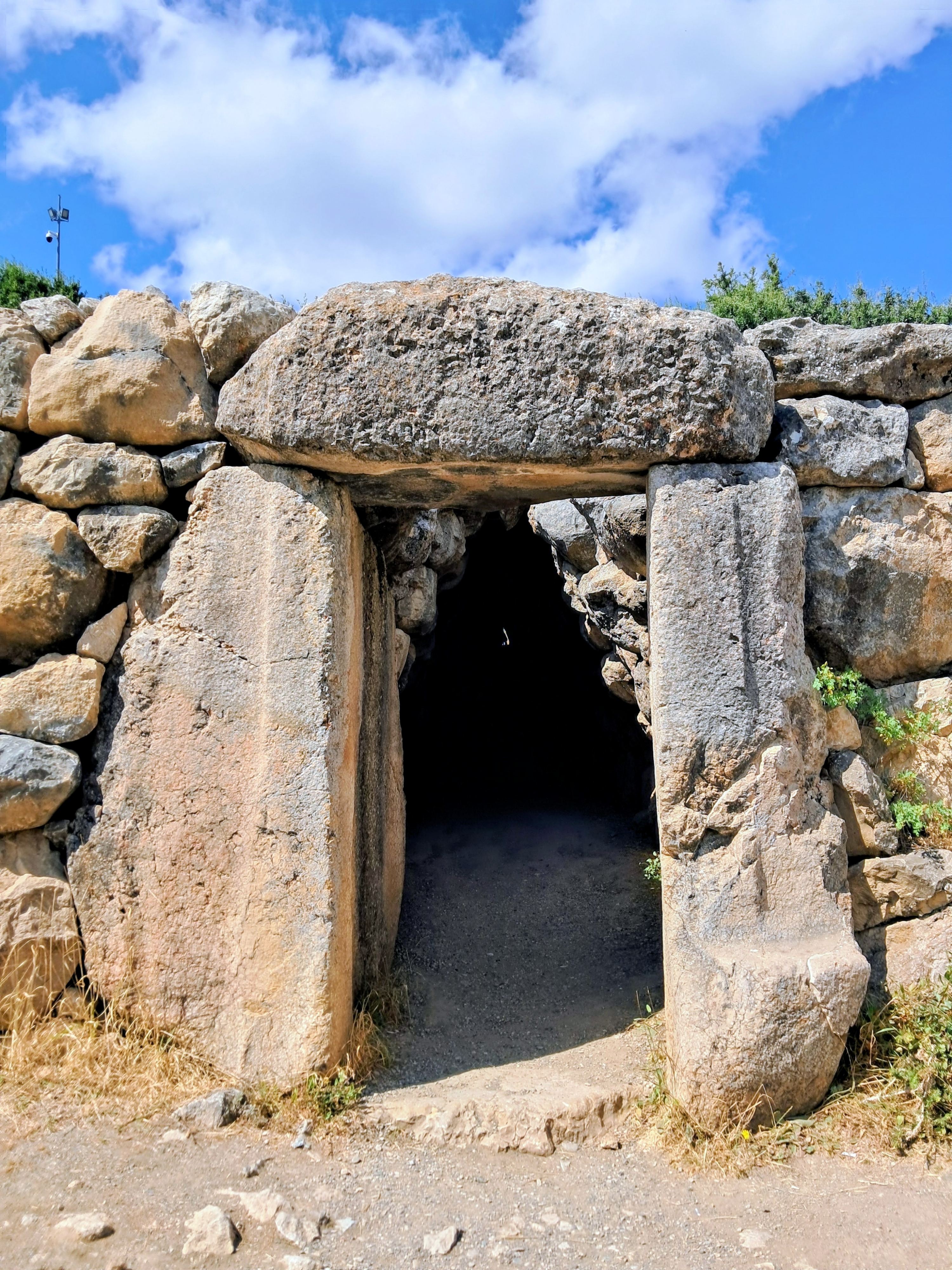 A stone doorway framed by large upright jambs and a heavy horizontal lintel opens into a dark underground corbelled tunnel beneath an ancient dry-stone rampart. The entrance leads into the shadows of the Yerkapı postern tunnel at the Hittite archaeological site of Hattusa in Turkey, with a faint pinprick of daylight visible in the far distance. Surrounding the entrance are weathered limestone blocks and patches of dry vegetation under the blue sky with scattered clouds.
