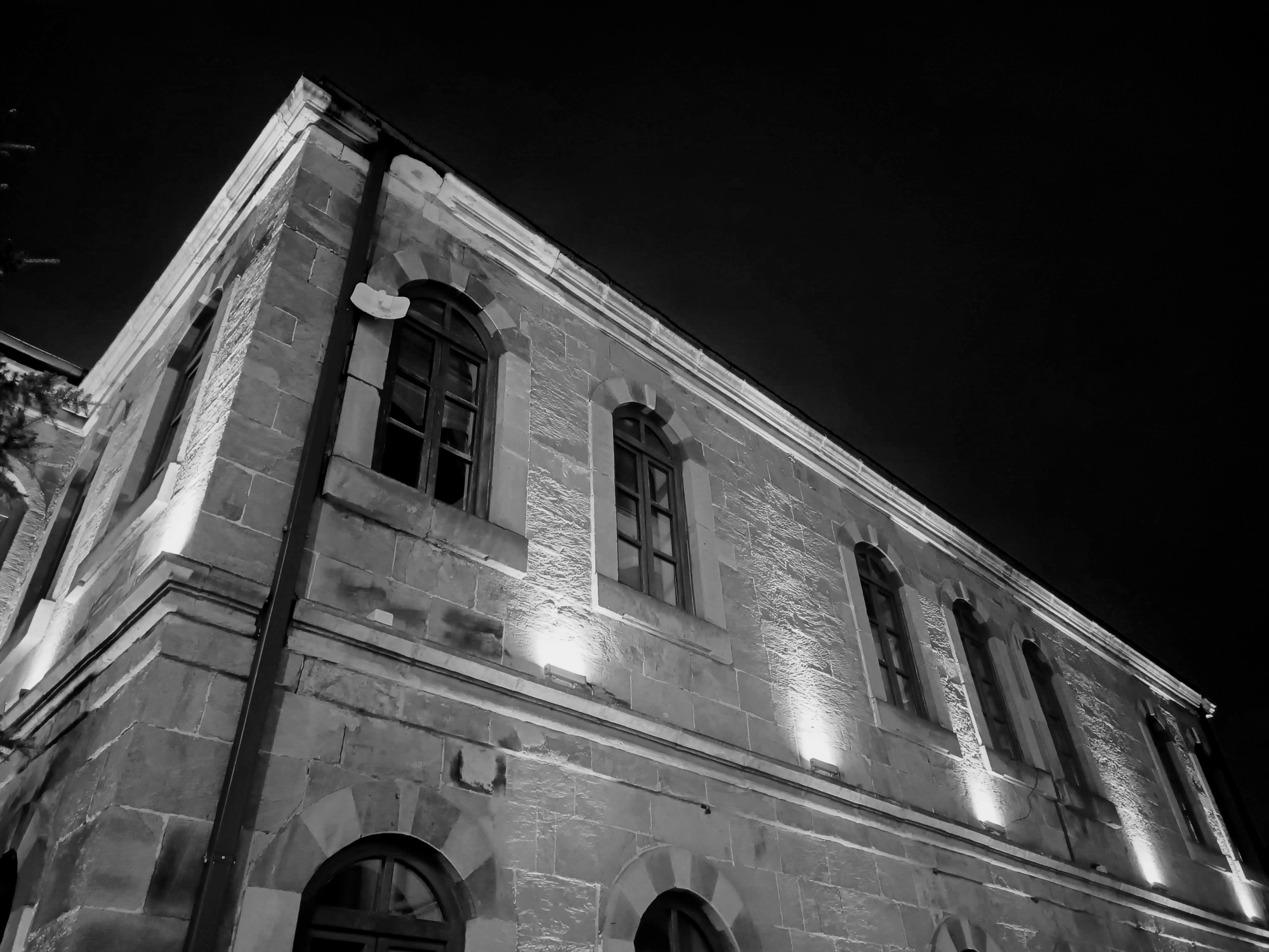 The side façade of a historical stone building illuminated at night. Upward-facing lights along the base showcase the textured masonry and series of arched windows lined up across the upper floor. Near the top left window, a small white fixture of some sort is attached to the wall. The building contrasts sharply against the dark night sky.