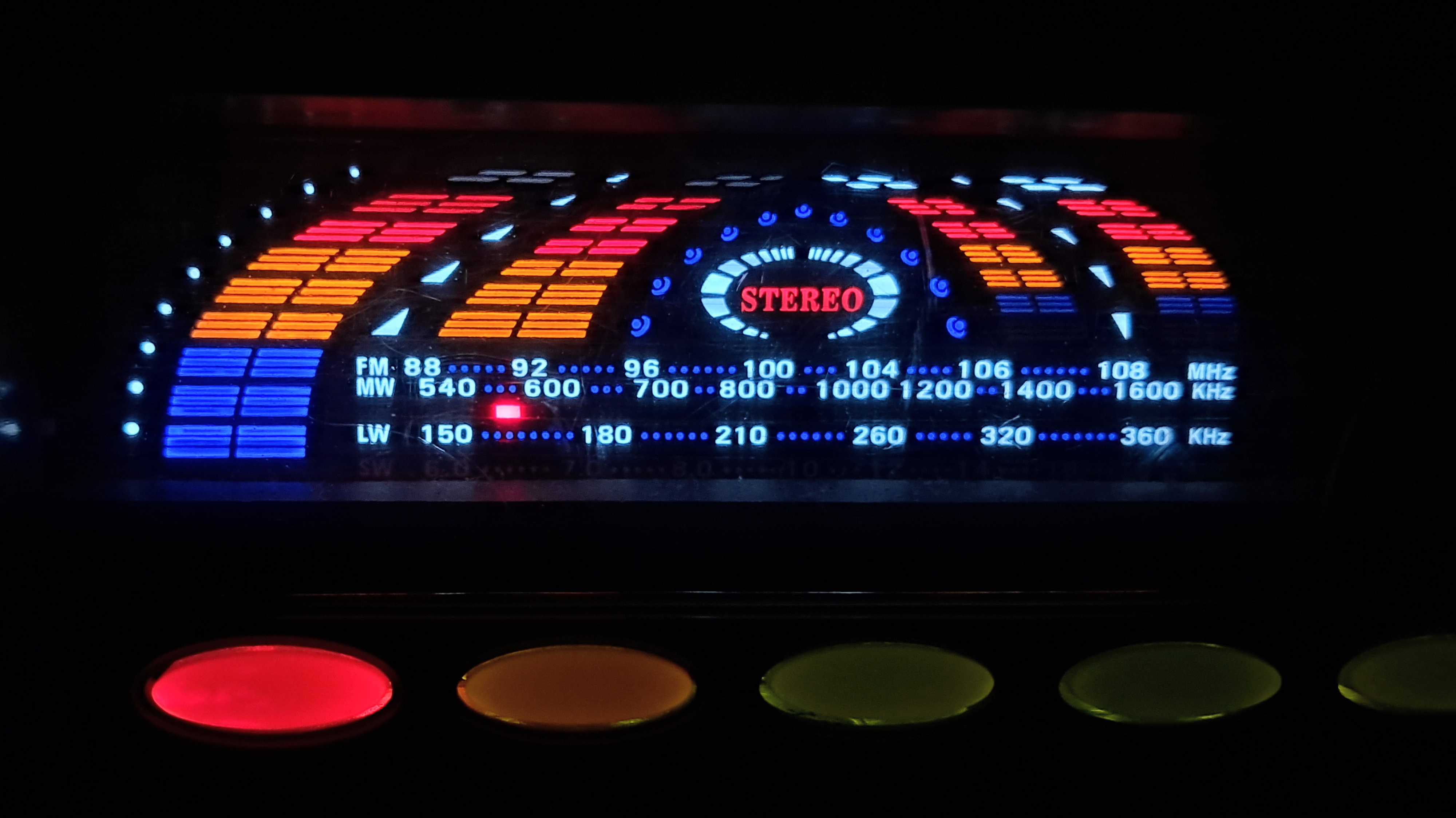 An illuminated vintage audio receiver display set against a dark background, featuring an arched graphic equalizer with curved bars lit in blue, orange, and red segments. In the center, a circular emblem glows with the word "STEREO" in bright red serif lettering. Beneath the equalizer, radio frequency scales are displayed in blue text: "FM 88 92 96 100 104 106 108 MHz", "MW 540 600 700 800 1000 1200 1400 1600 KHz" marked by a small red tuner indicator, and "LW 150 180 210 260 320 360 KHz", along with a faint bottom row to indicate shortwave frequencies. Along the bottom edge sits a horizontal row of rounded buttons backlit in red, amber, and dark green.