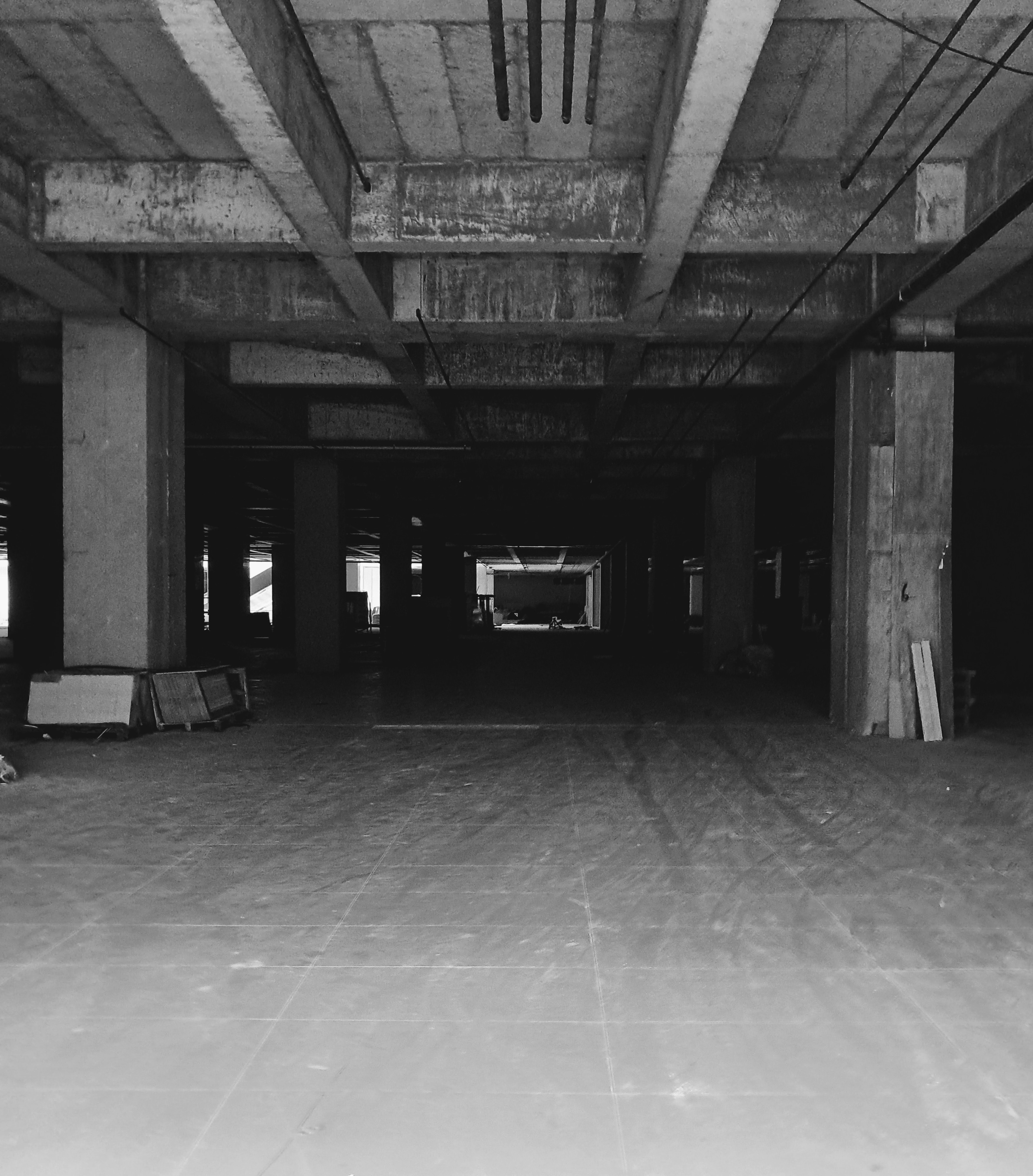 A view looking down the length of an unfinished concrete building interior. Heavy concrete support beams run across the exposed ceiling with hanging pipes, supported by square pillars along the sides. In the foreground, the concrete floor shows faint scuffs and tire tracks, while stacked building materials lean against the pillars. The deep interior recedes into heavy shadow, with some light in the center distance.