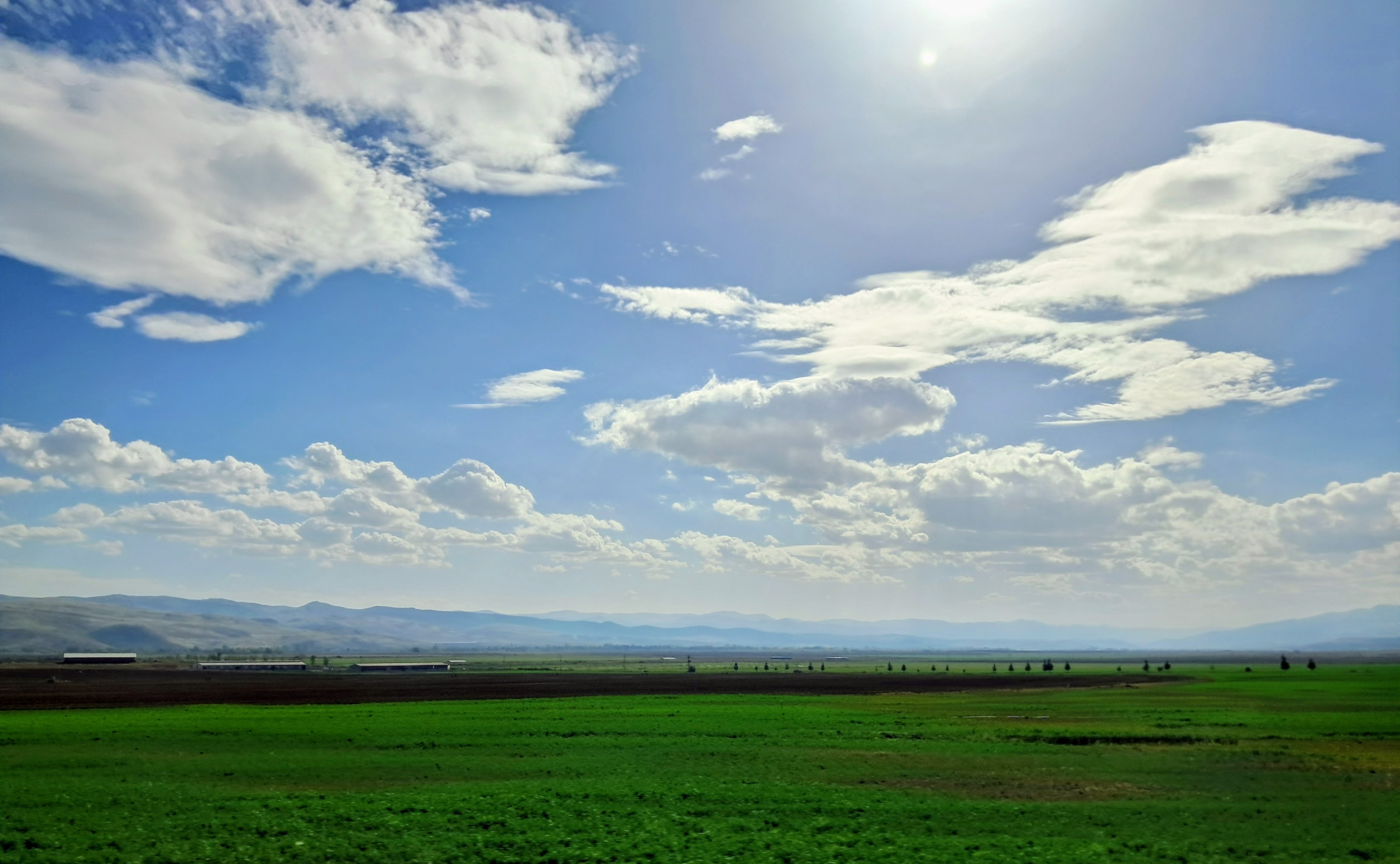 A landscape featuring a green field under a blue sky filled with scattered white clouds. The foreground consists of a vibrant green agricultural field that borders a dark brown patch of bare earth. Hazy rolling blue mountains span the distant horizon beneath the bright sun. On the left side of the fields are some low, flat structures that are long agricultural sheds. A row of tiny silhouettes of distant trees are also visible.