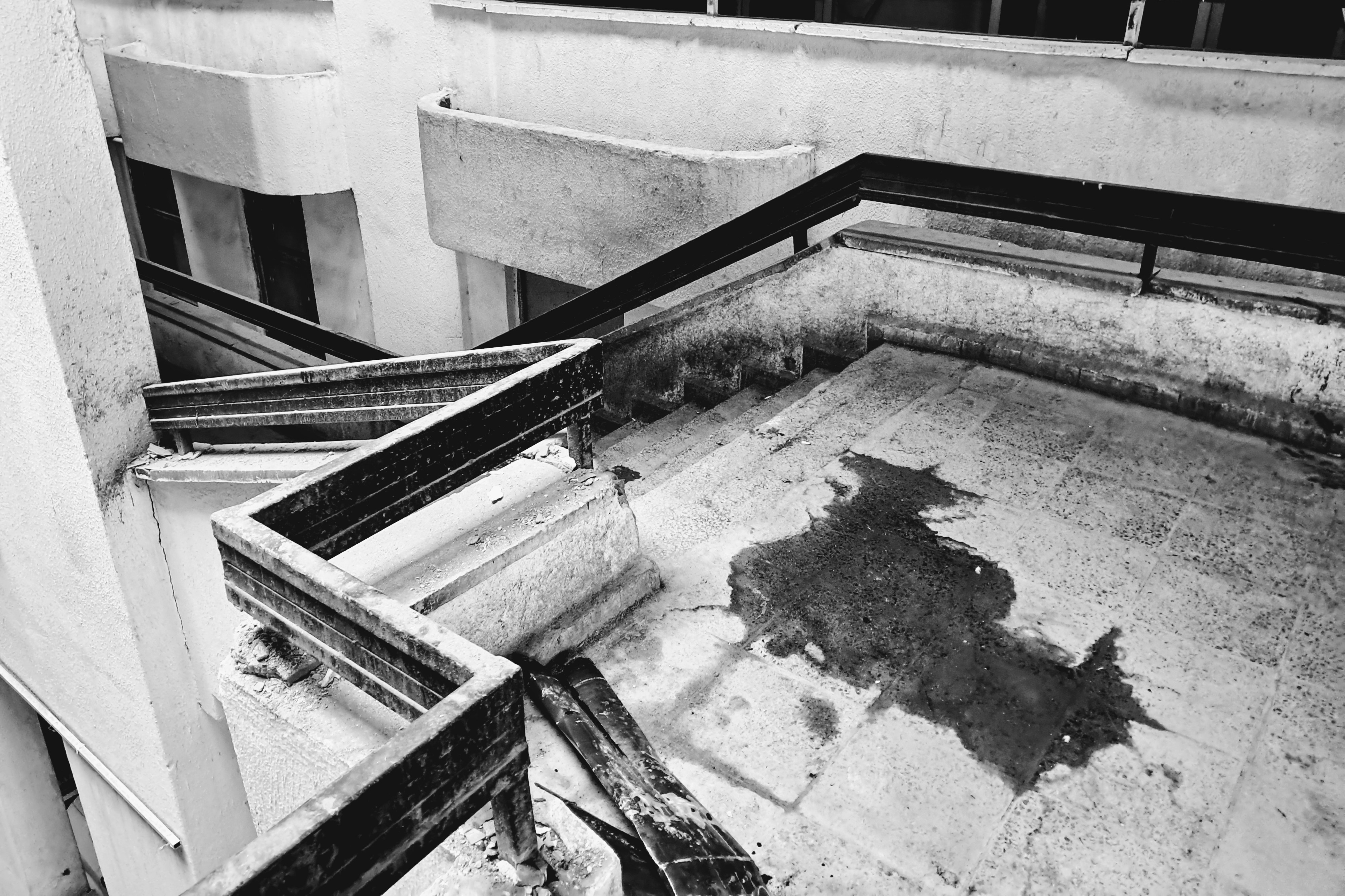 The weathered stairwell and landing of a multi-story concrete building. Angular dark metal railings are placed alongside worn steps that lead down to a tiled terrace. A large dark puddle sits on the pavement, accompanied by scattered debris and peeling material near the railing base. In the background, geometric concrete facade elements and window openings appear.