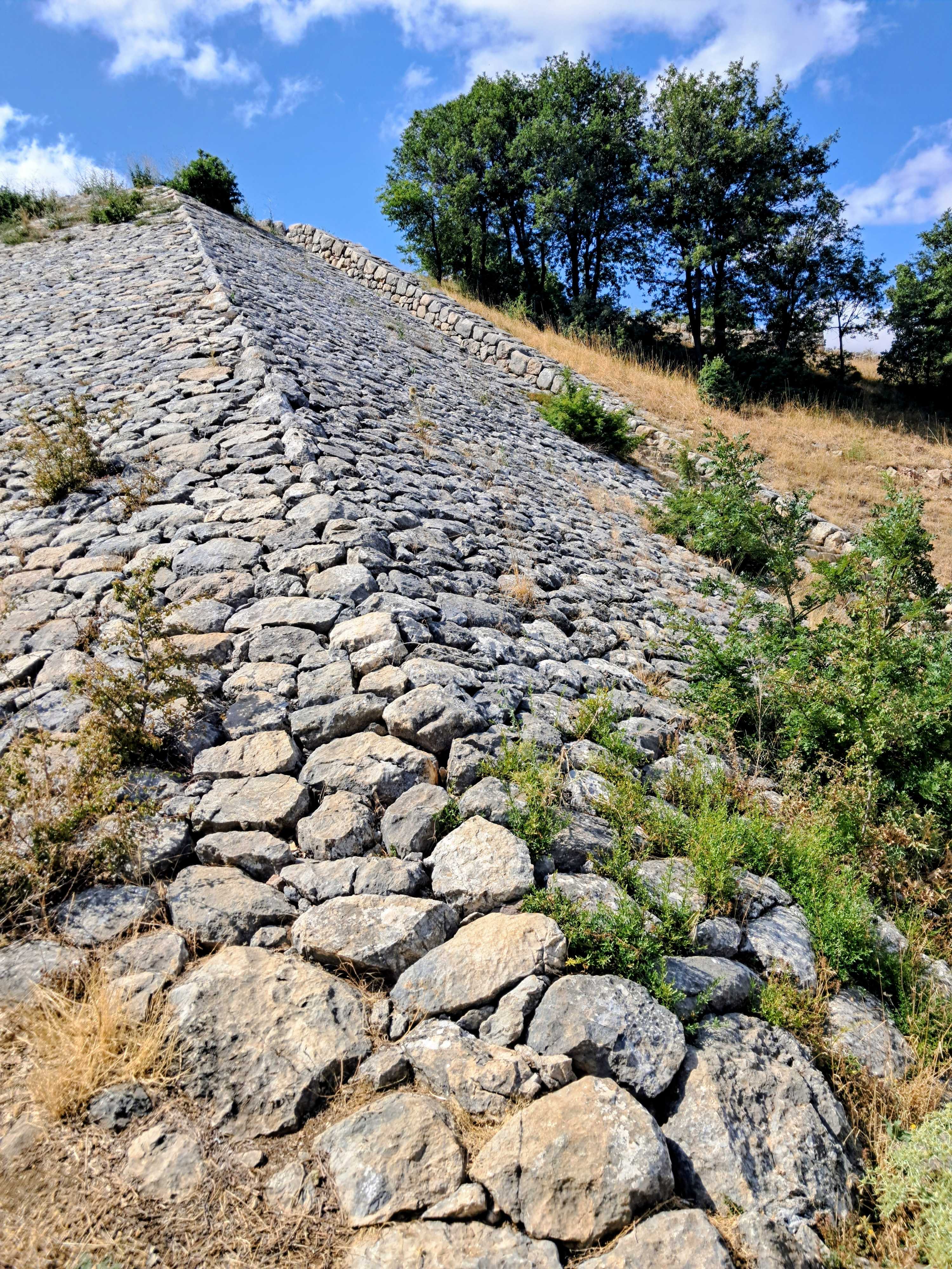 A steep ancient stone rampart constructed from rough-hewn gray boulders rises diagonally up a hillside. Patches of green shrubs and dry yellow grass sprout along the rocky surface and the adjacent slope. At the top of the incline, a row of lush green trees stands against a blue sky with scattered white clouds.