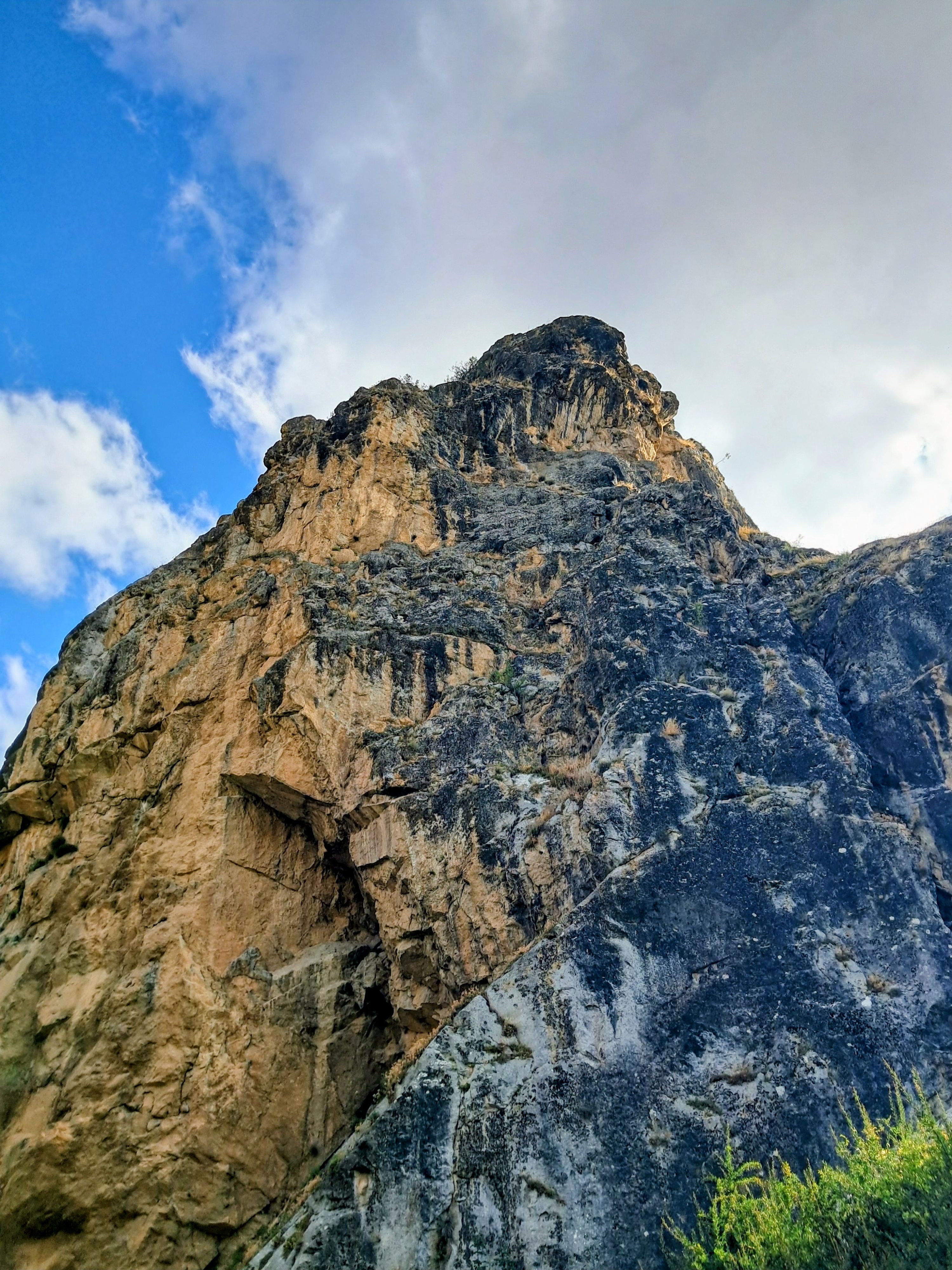 Wide view of rugged rock formation against blue sky with white clouds, showing textured brown and dark gray stone surfaces with sparse green grass at bottom right.