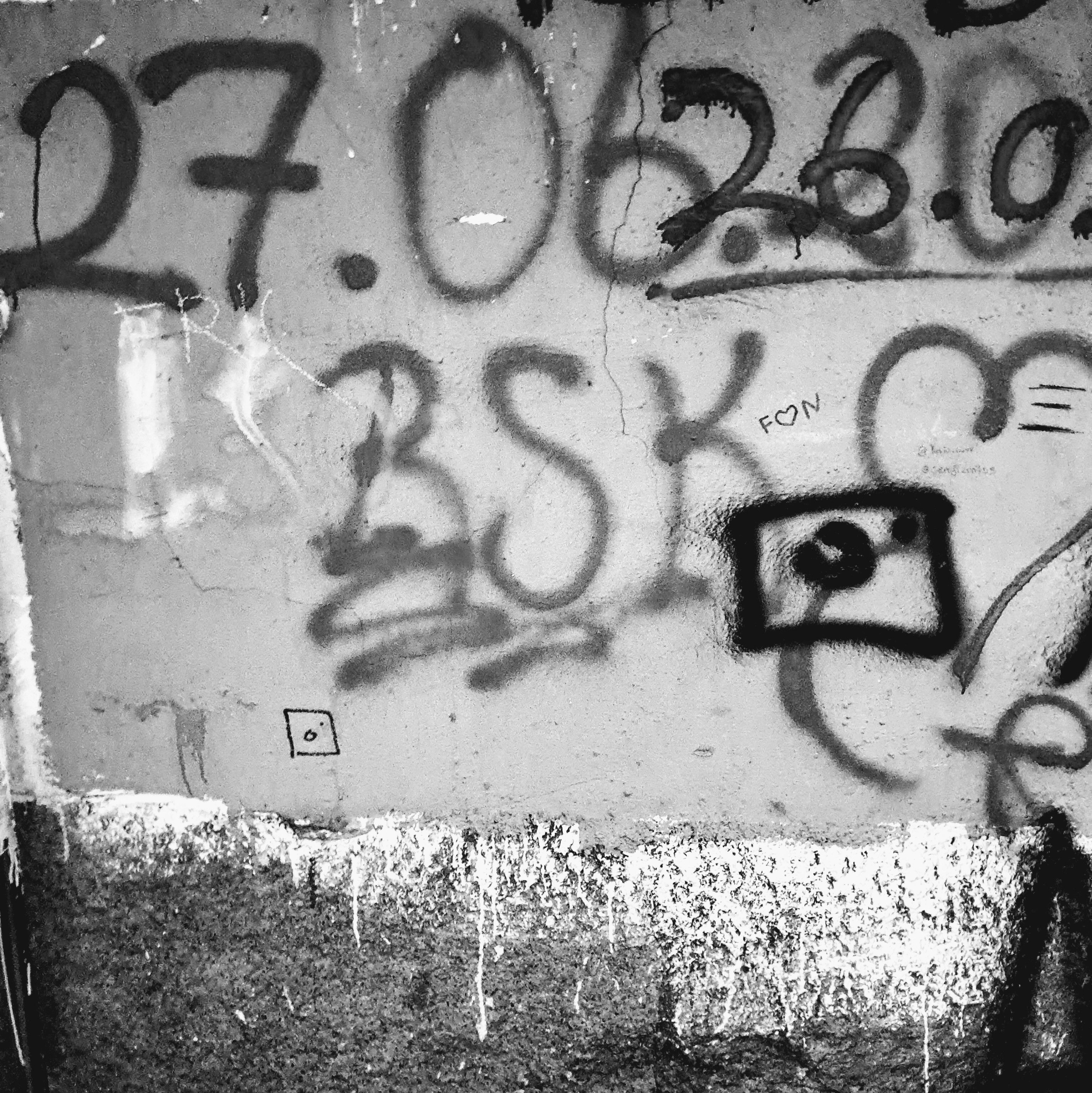 A weathered concrete wall is covered in black spray-painted graffiti and various hand-drawn markings. Across the top, large numbers read "27.06", "26.02" and similar date formats. Below the numbers, stylized graffiti letters spell out "BSK", positioned in the middle of multiple doodles resembling the Instagram® logo, a heart outline, and the text "F♡N". The bottom portion of the view shows a rough concrete part with white paint splattered across it.