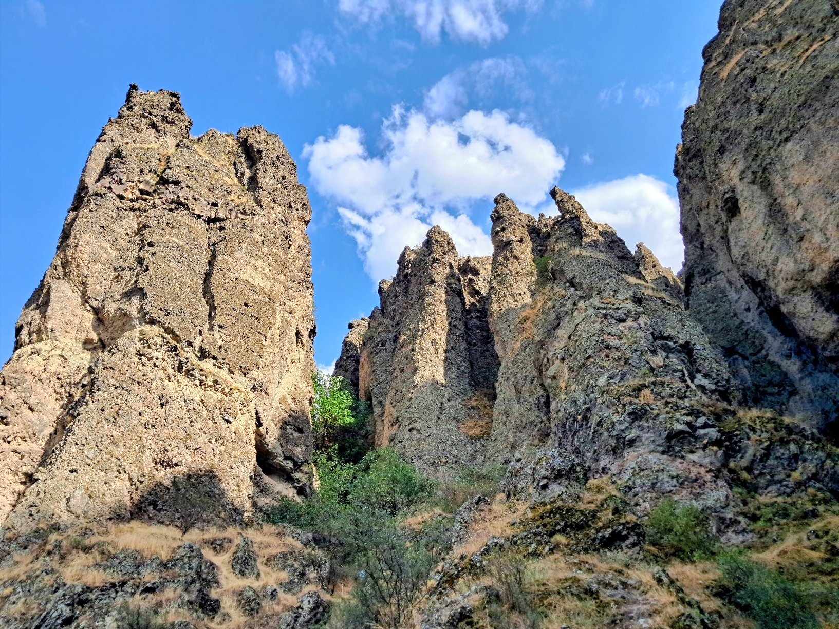 A low-angle view looking up at tall jagged rock spires and cliffs made of weathered brown stone. Sparse green trees and patches of dry grass are stuck to the base and crevices between the formations, all under the blue sky with soft white clouds.