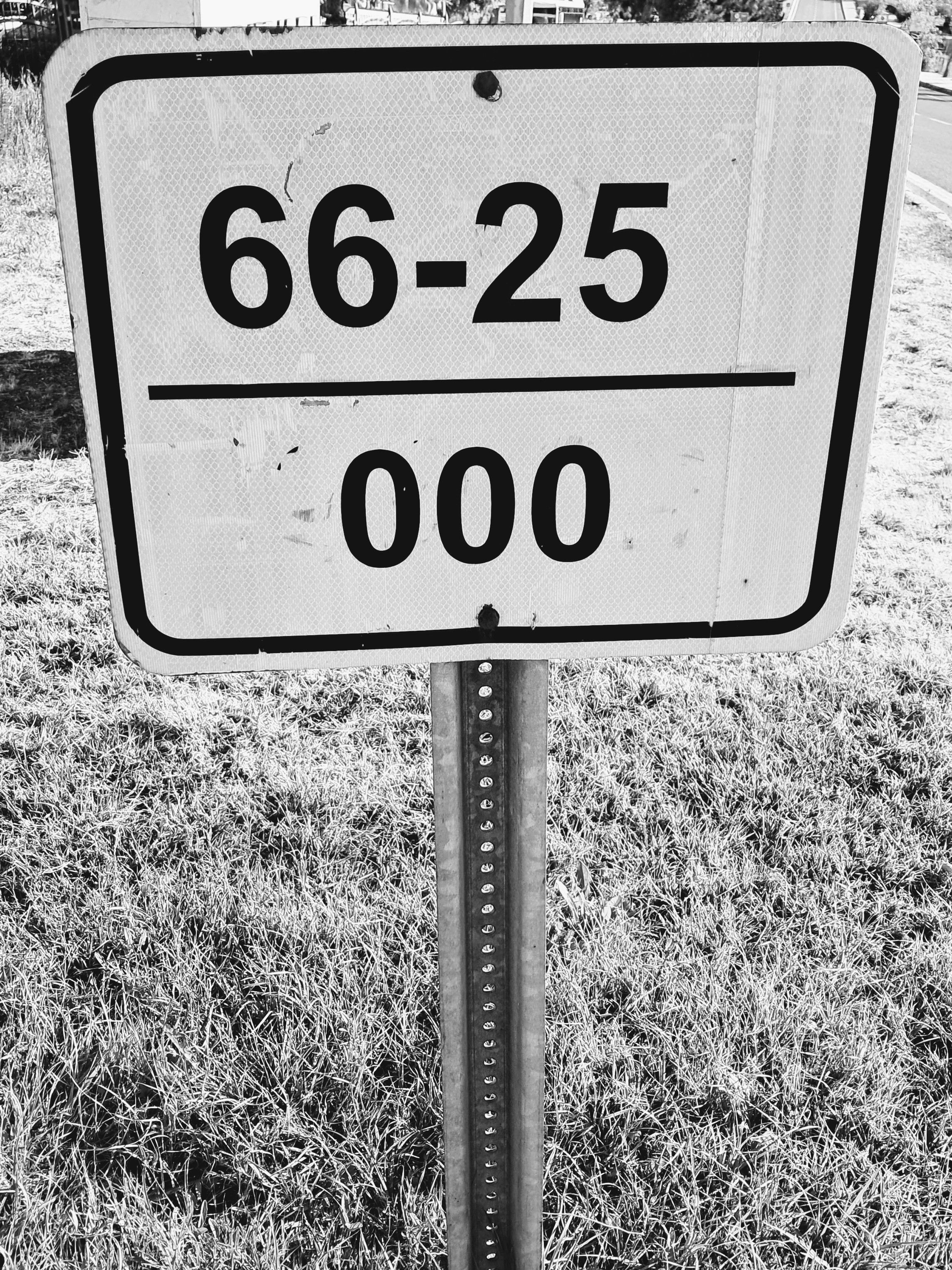 Close-up view of a rectangular metal reference marker road sign with rounded corners mounted on a metal post above a grassy patch. The sign features a black inner border and is divided horizontally by a black line, displaying the numbers "66-25" on the top half and "000" on the bottom half.