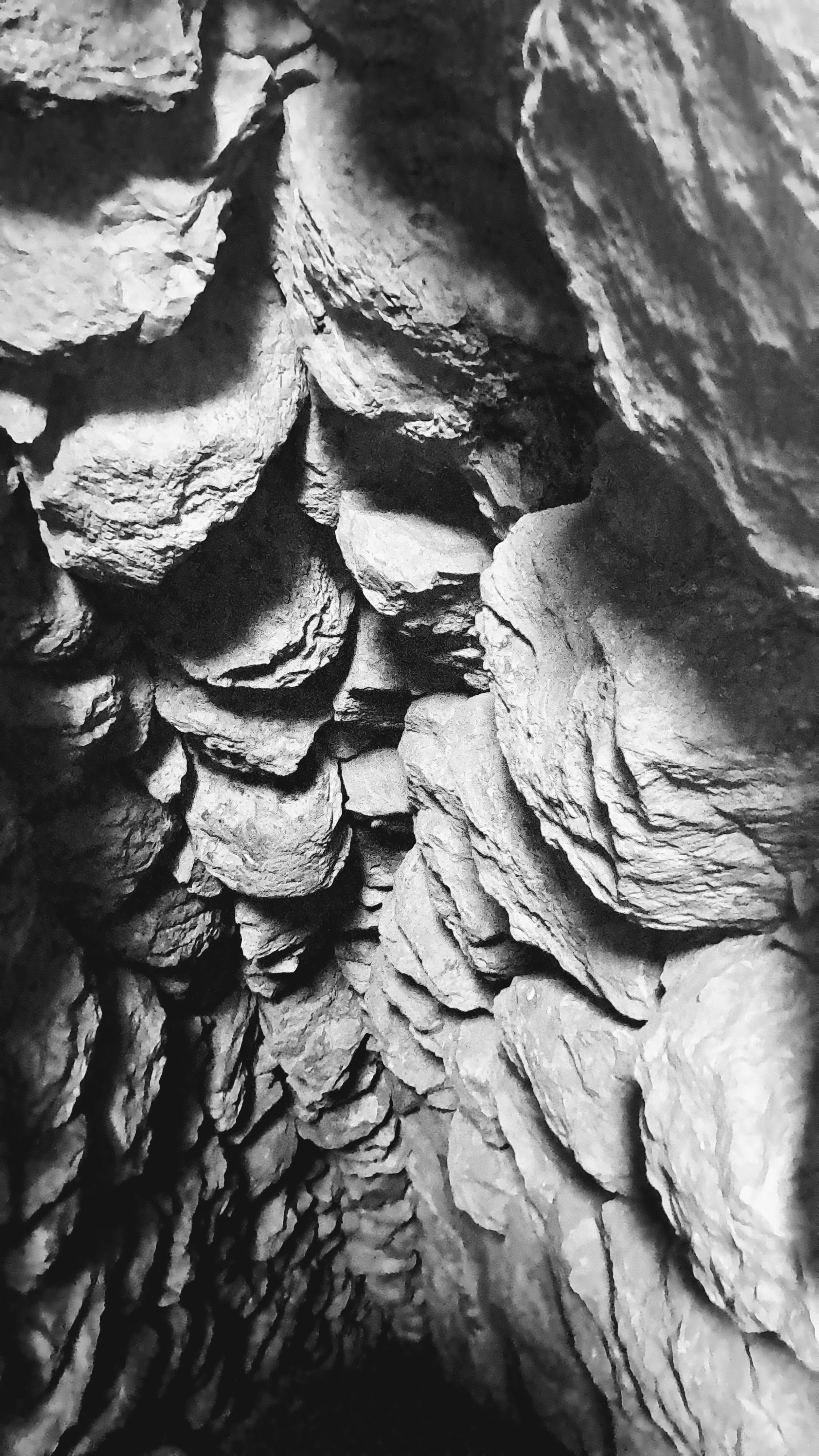 A view looking down into a narrow rugged stone shaft. Rough and fractured rocks with sharp edges and heavy textures cover the tight enclosure, with light behind hitting the upper ridges and casting deep shadows into the dark abyss below.