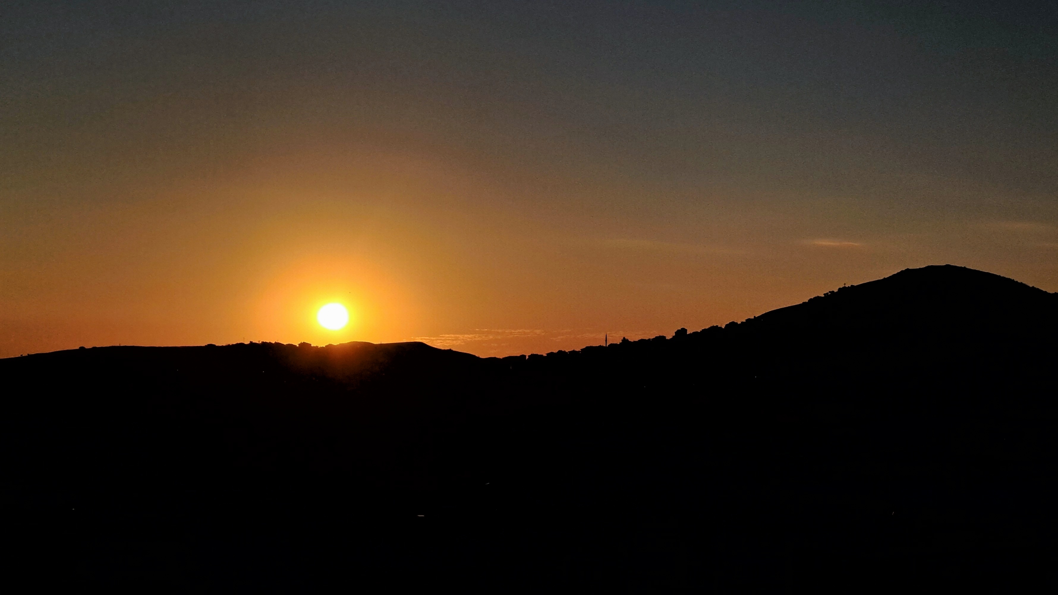 A view of sunset featuring the bright golden sun low along the horizon casting an orange and yellow glow across the clear sky that fades into deep blue at the top. The foreground consists entirely of a dark silhouette of rolling hills and a mountain peak on the right. Tiny indistinct buildings and city structures along the ridge line are all silhouetted against the vibrant sunset light.