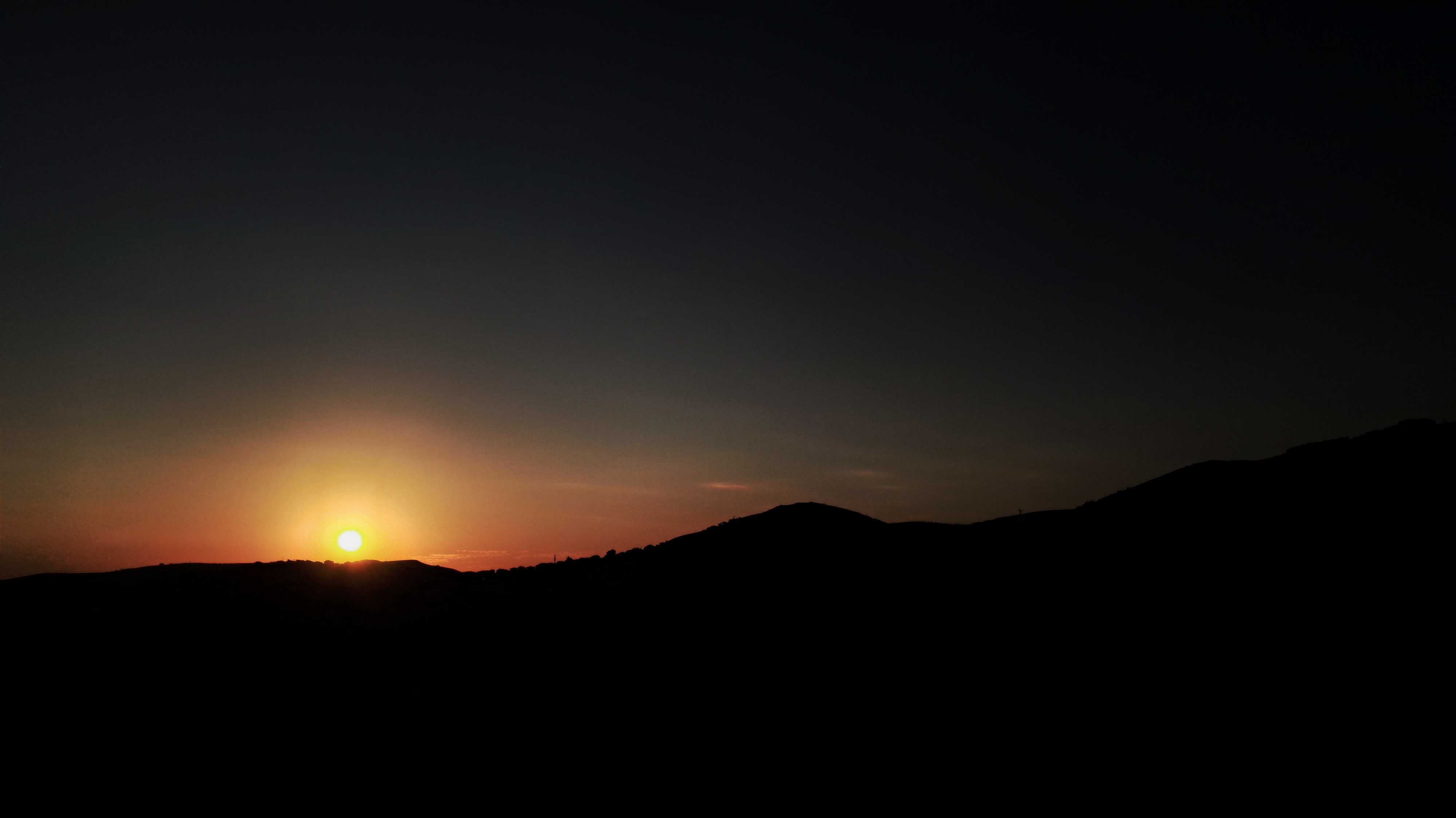 View of bright sun low on horizon behind silhouetted mountain ridgeline, with gradient of orange light fading into dark black sky.