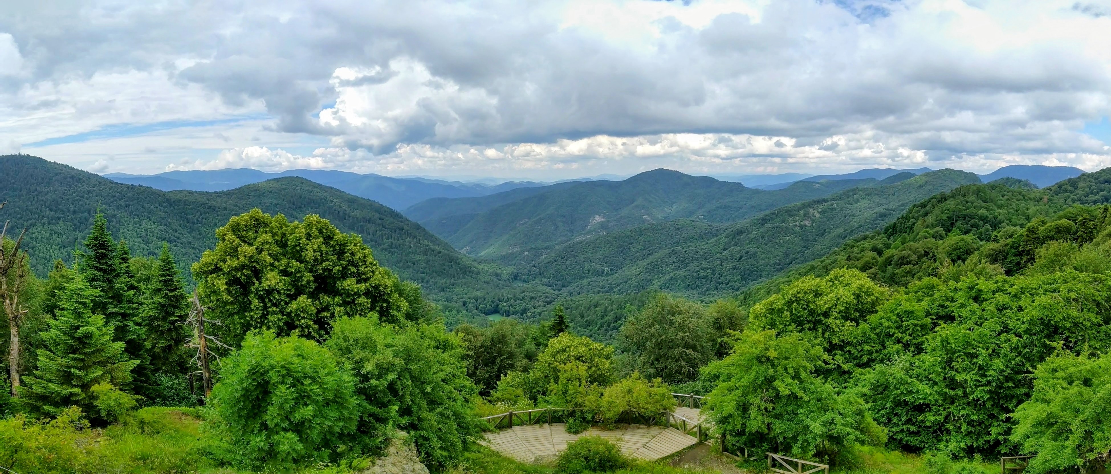 Oanoramic view looking out over densely forested green mountain ridges under a cloudy sky. In the foreground, deciduous and evergreen trees fill the viewing area featuring wooden decking bordered by railings. The rolling mountain ranges stretch into the distance, gradually fading into hazy blue hues beneath the gray clouds.