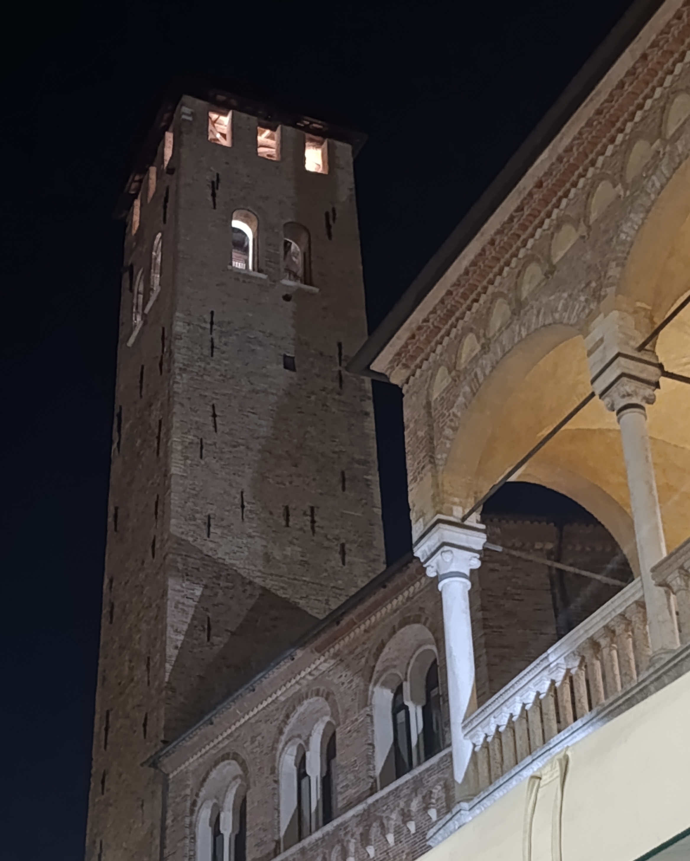 Torre degli Anziani e un angolo di Palazzo della Ragione visti dal basso, illuminati. Foto scattata di notte.
