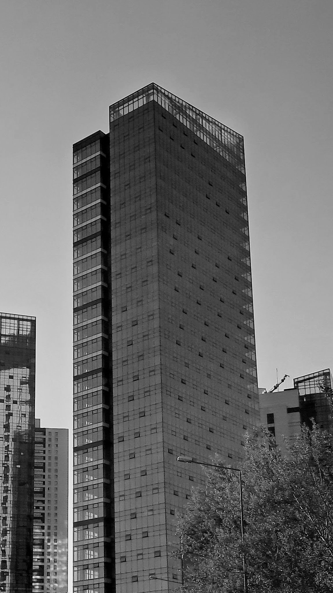 Urban area featuring a modern high-rise skyscraper towering against the blue sky. The building features a dark façade with a repeating grid pattern of windows and small square openings across its front, flanked by a glass-paneled corner structure and topped with a glass-enclosed level. Portions of other contemporary buildings appear on either side, while a streetlamp and lush tree foliage are on the lower foreground.