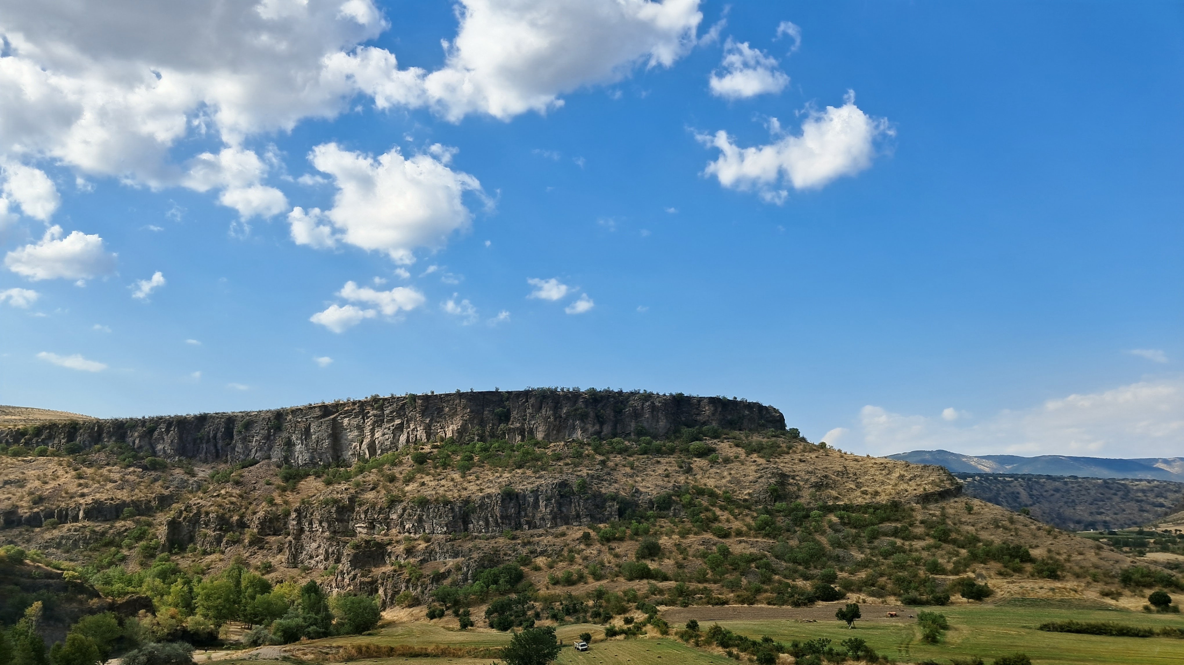A view of a wide mesa featuring a prominent flat-topped rocky plateau with steep cliffs under a blue sky with scattered white clouds. The rugged terrain slopes down into a valley filled with a mix of green trees, shrubs, and dry brush. In the lower valley area, there is a truck and a bunch of hay bales. Distant rolling hills are visible in the background to the right.