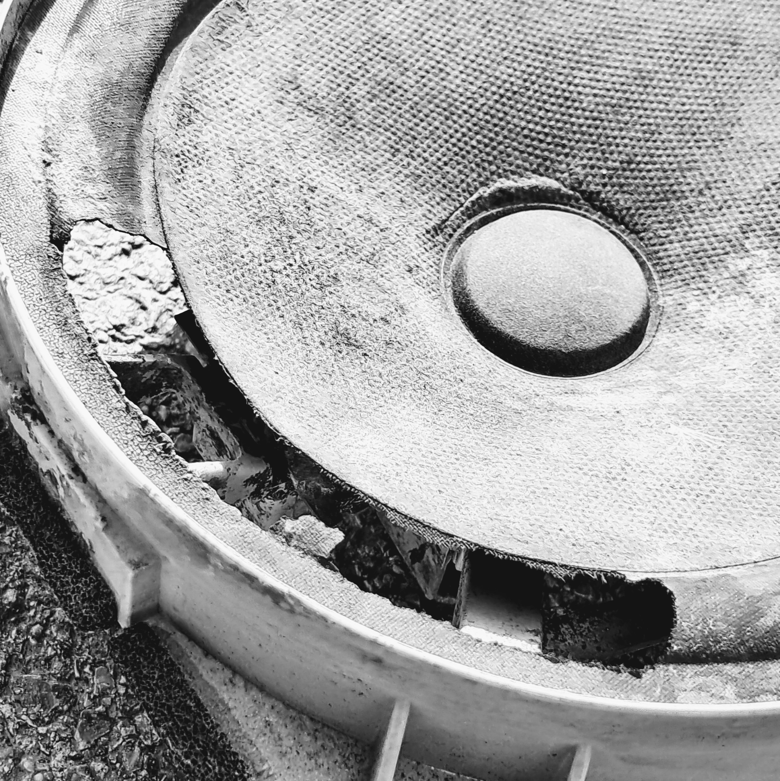 Close-up view of a severely damaged loudspeaker driver set inside a plastic mounting frame. The outer rubber suspension surround has rotted away and began to torn apart along the lower-left edge, exposing the internal basket and frame supports. The main textured cone and central dust cap remain largely intact, resting on an uneven asphalt ground surface.