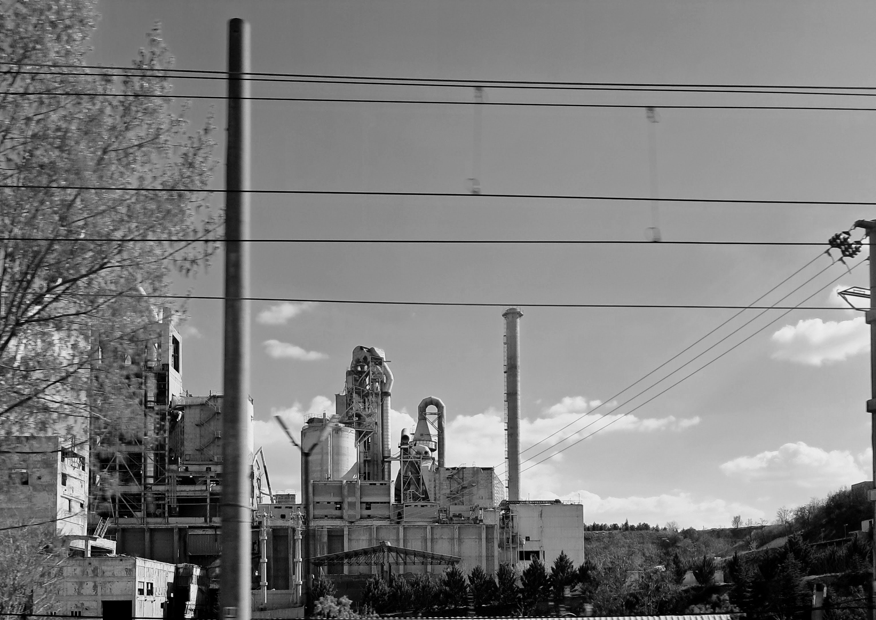 A sprawling industrial complex with multiple structures, silos, and a tall smokestack seen through commuter train. The foreground includes horizontal power lines and a vertical pole of some sorts. Trees show up on the left side and bottom edge of the frame beneath a blue sky with scattered clouds. The exact type of facility is uncertain, but it appears to be a cement plant or some sort of manufacturing site.