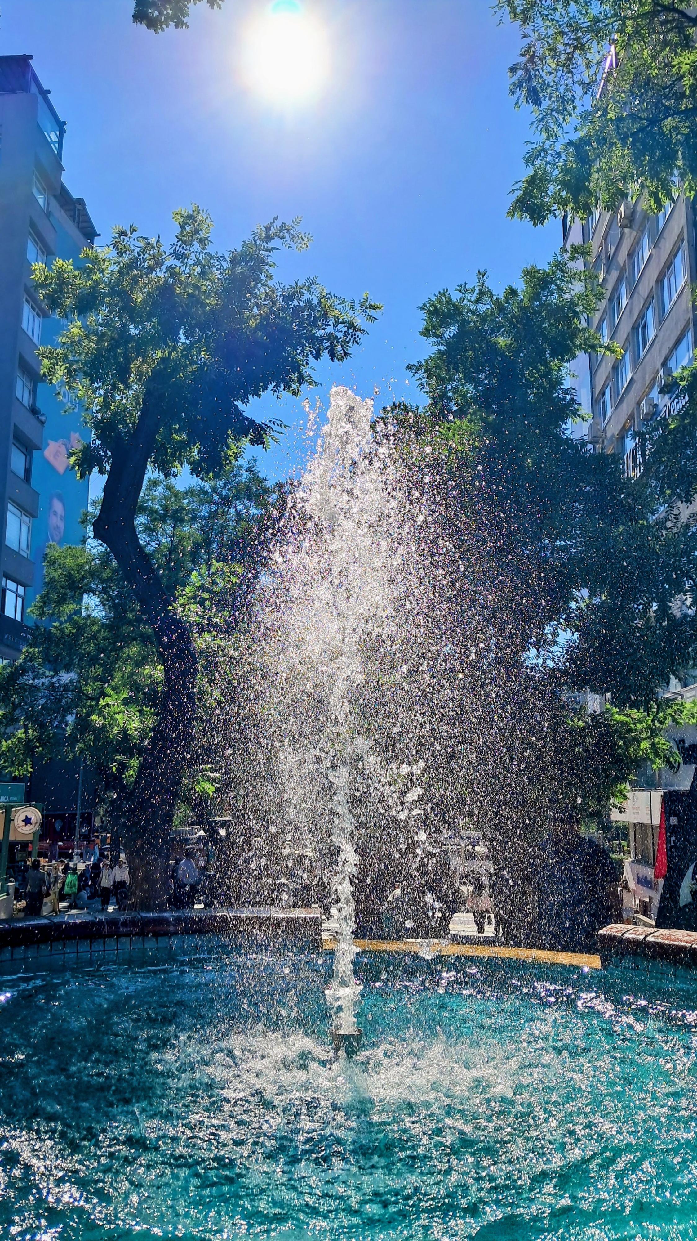 A central water fountain shoots a high spray into the air, its sparkling droplets illuminated by bright sunlight under a clear blue sky. The turquoise circular stone basin below is filled with shimmering clear water. Surrounding the fountain are lush green leafy trees, framed by multi-story city buildings on either side.