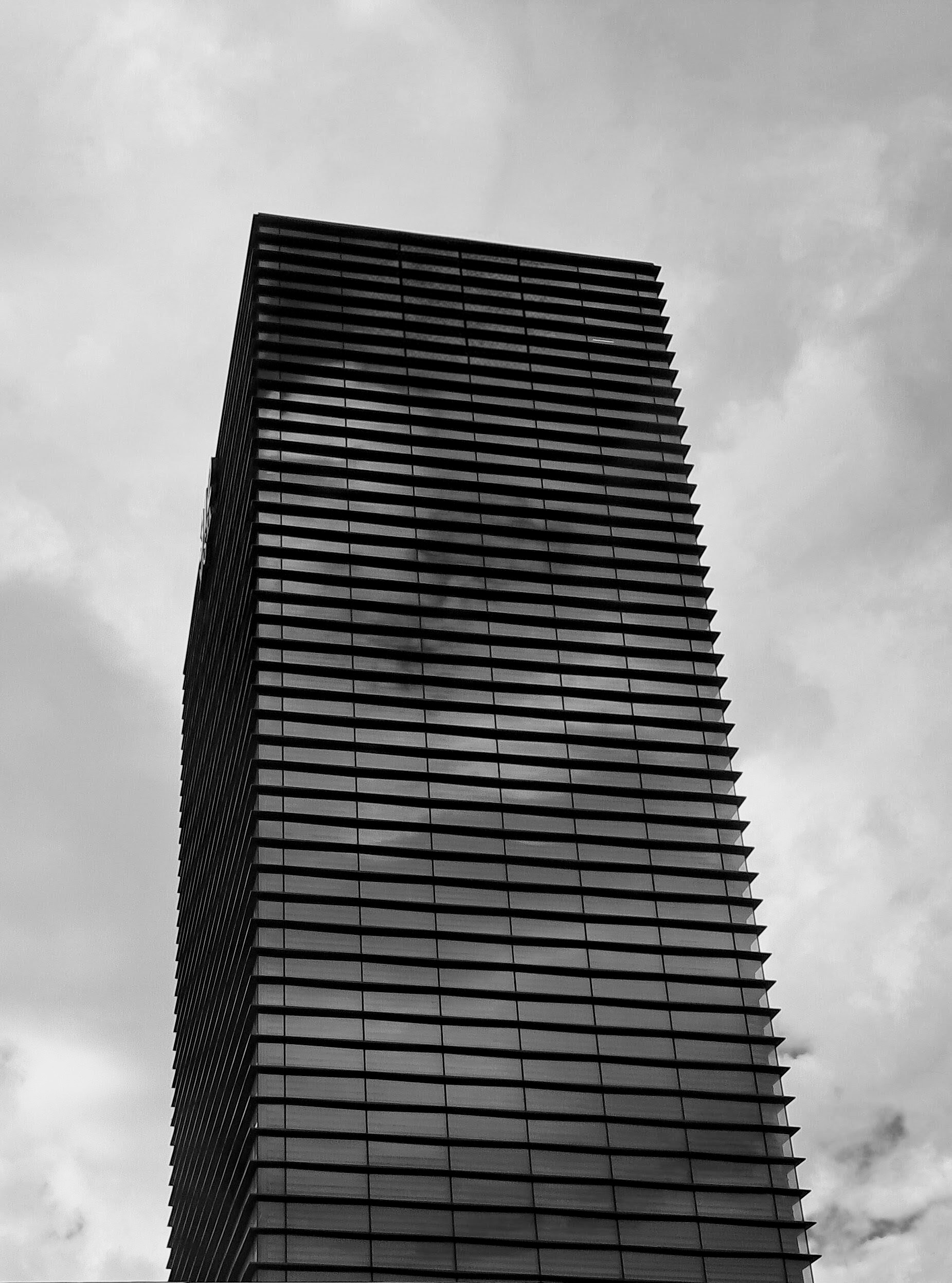 A modern skyscraper rising against an overcast sky. The façade features closely spaced horizontal architectural fins running up the entire height of the building for that dense ribbed texture over the glass exterior.