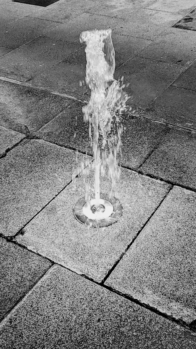 A vertical stream of water shoots upward from a circular metal jet embedded in a paved stone walkway, highlighting the splashes and movement of the fountain against the square textured stone tiles.