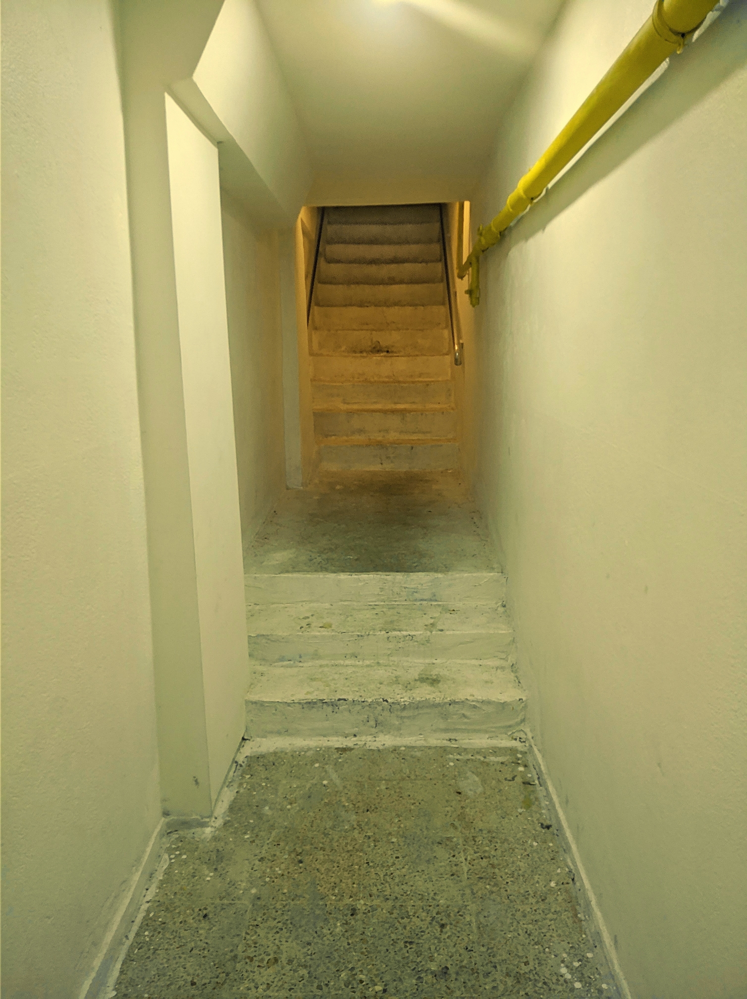 A narrow dimly lit indoor hallway with plain off-white walls leads toward concrete stairs ascending in the background. A bright yellow pipe runs along the upper right wall. The floor in the foreground consists of speckled terrazzo tiles leading up to a short set of white concrete steps.