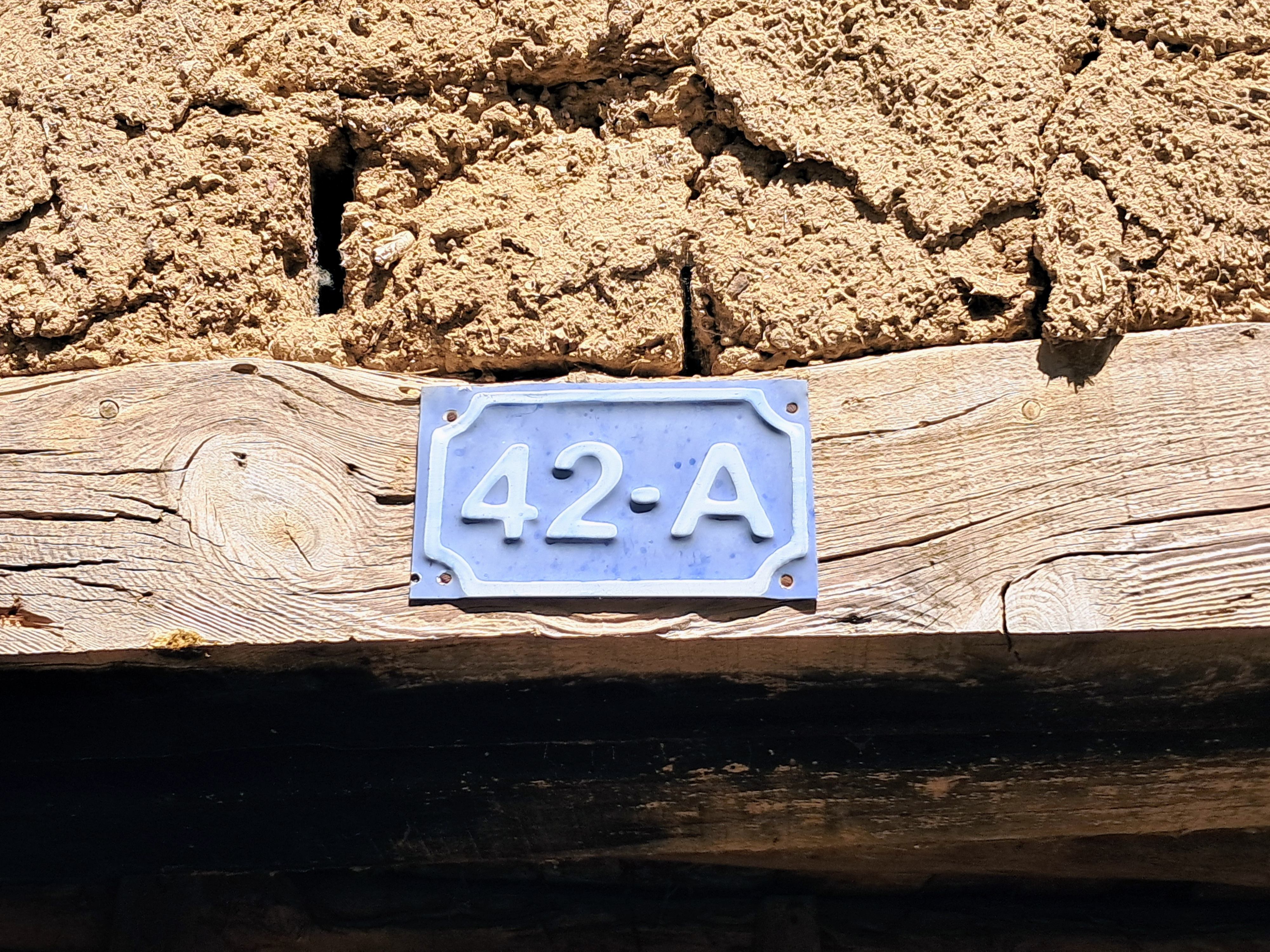 A light blue rectangular sign made of faded metal displays the white text 42-A and is attached to a weathered wooden beam. Above the wood is a rough wall made of yellow cracked dried earth, and below the beam is a dark shadowed gap into the deep dark...