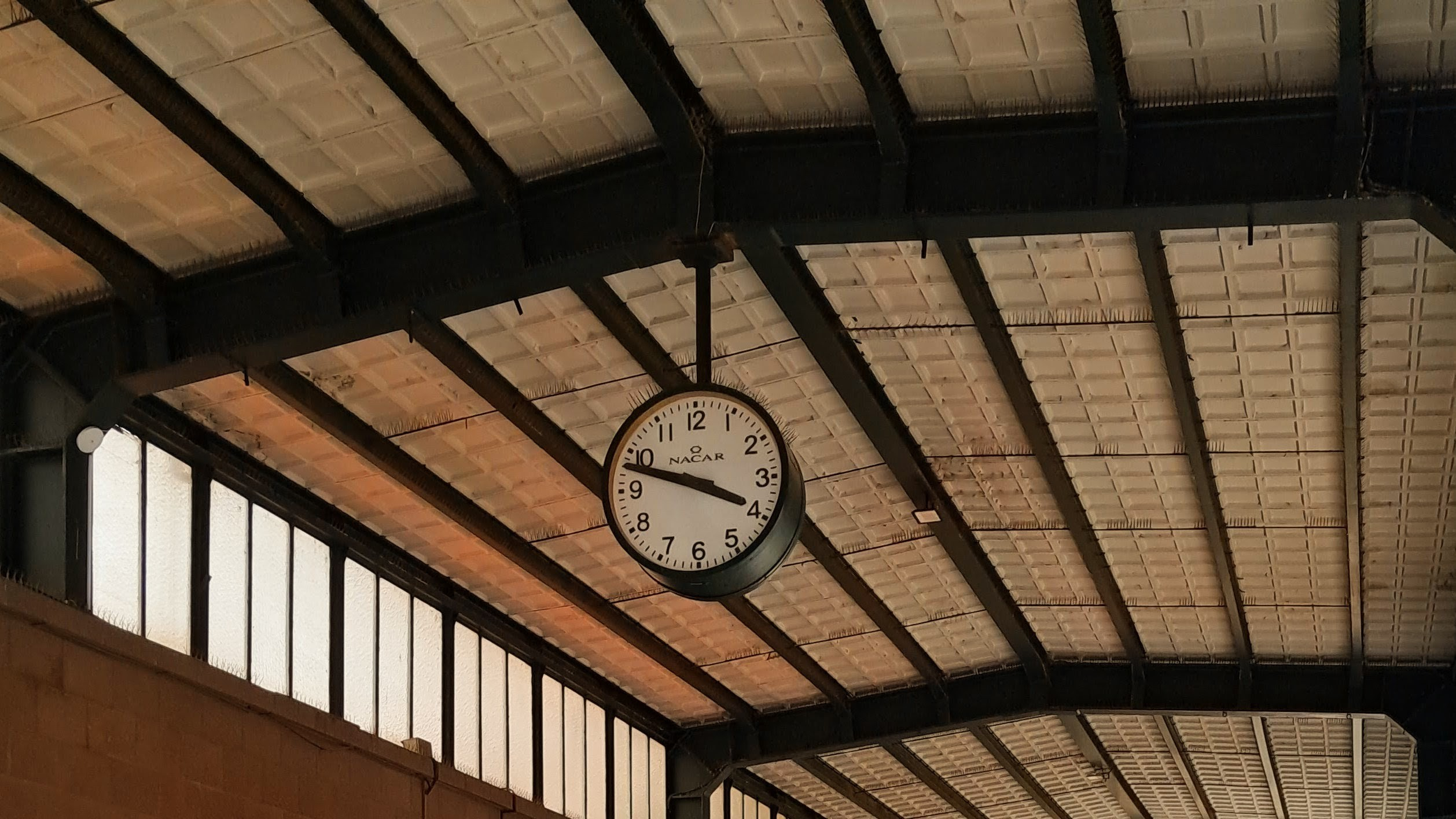 A round analog clock with black Arabic numerals against a white face hangs suspended from dark metal beams beneath an industrial vaulted ceiling inside a train station. The clock hands are positioned at 4:47. Behind the clock, angled architectural beams support rows of textured ceiling tiles, illuminated by natural light appearing through a row of vertical frosted glass window panes along the left wall.