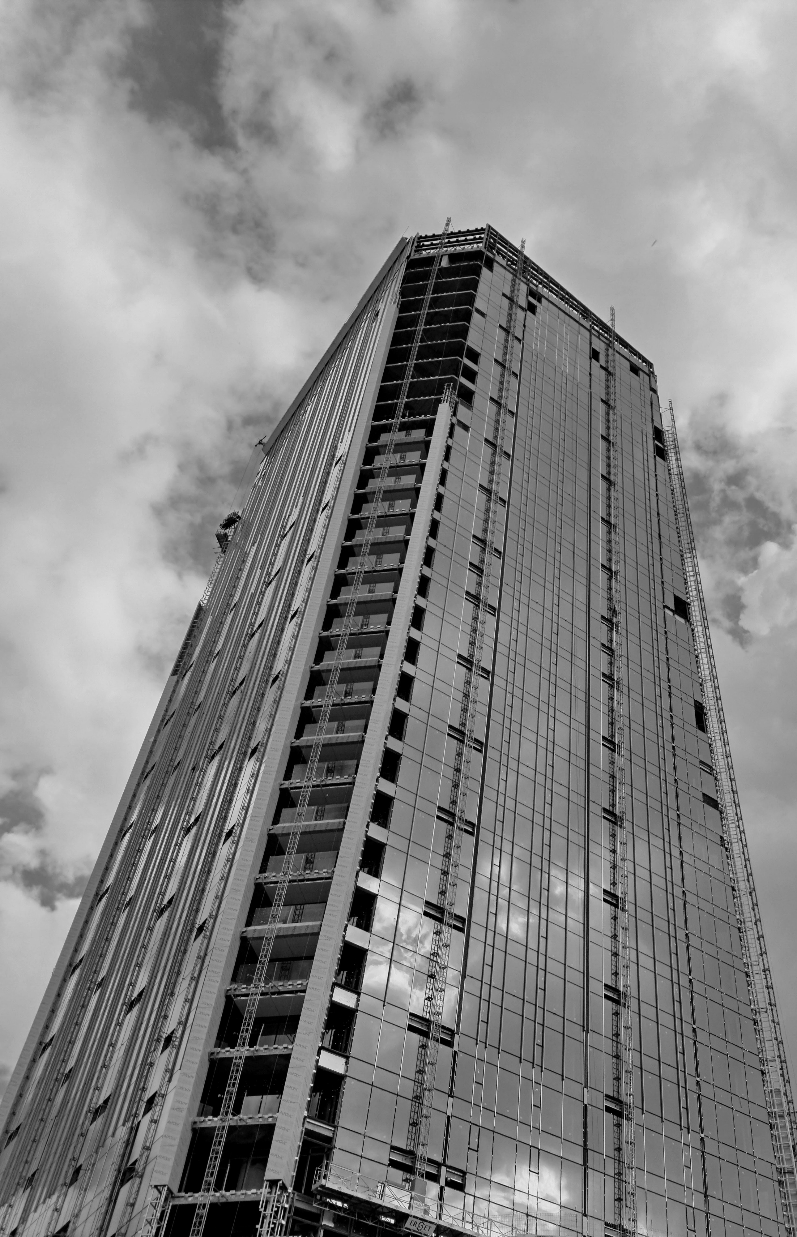 A modern skyscraper under construction against a blue sky with scattered clouds. The high-rise building features extensive reflective glass façade panels mirroring the sky, alongside sections of exposed concrete framing and unfinished balconies. Vertical construction hoist tracks run along the exterior walls, with a suspended work platform near the lower section.