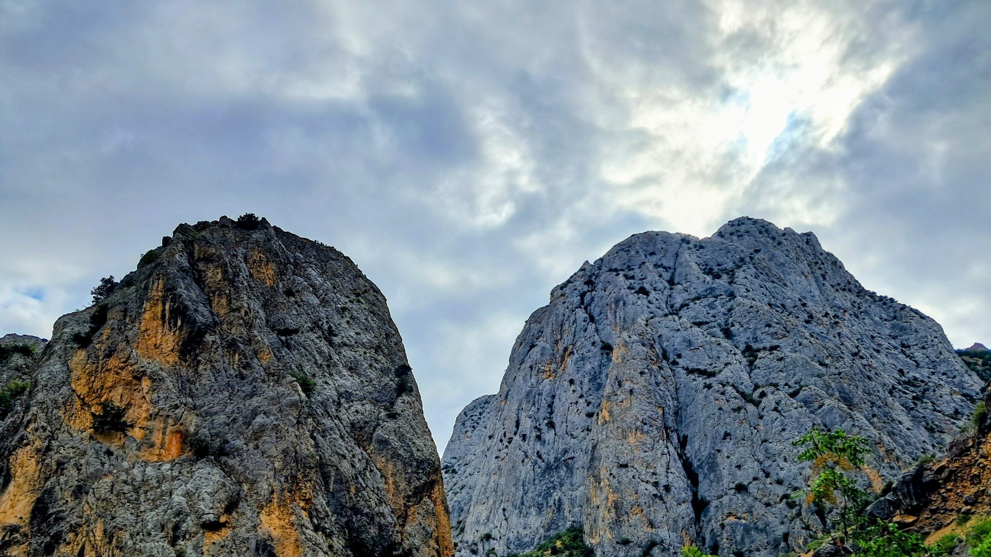 Two massive rugged rocky mountain peaks rise steeply against an overcast sky illuminated with bright sunlight filtering through the clouds. The rock face on the left features craggy weathered stone with patches of orange-tan color and sparse green shrubs clinging to the crevices. To the right, a larger craggy gray mountain peak fills the the frame with heavily textured fractured rock surfaces, framed by scattered green foliage along the bottom edge.