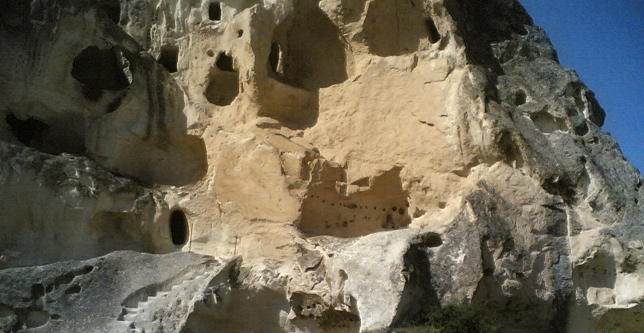 A massive light colored stone cliff face contains multiple dark hollowed out cave entrances and "windows" carved into the rock. Stone steps are visible near the bottom left, leading up the formation, while a clear blue sky is visible in the upper right corner. The structure is an ancient rock cut dwelling in Cappadocia.