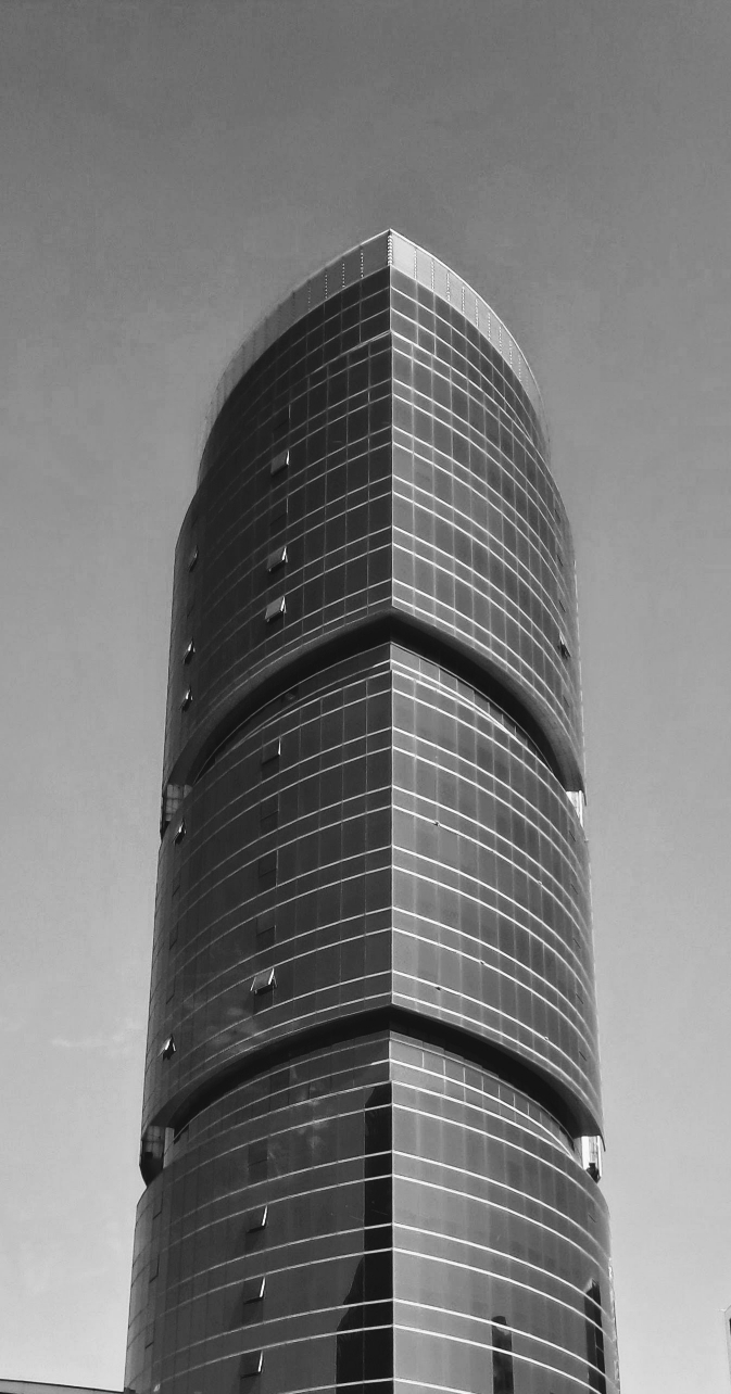 A tall curved glass skyscraper reaching into a clear blue sky. The building features a reflective façade that is divided into sections by thick horizontal gaps. The glass panels mirror the surrounding buildings.