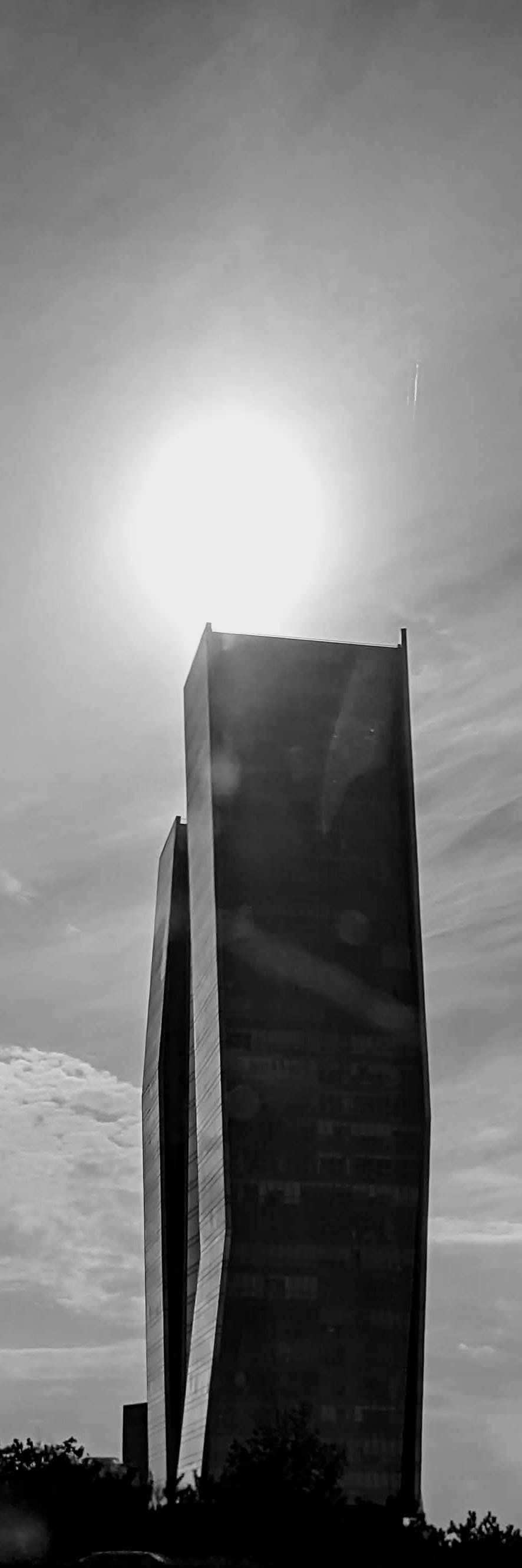 A tall modern skyscraper with angular glass façade rises from behind tree silhouettes against a hazy blue sky in the morning. The bright sun glows intensely right above the top edge of the tower, casting faint lens flare reflections along the building surface. A faint vertical streak appears in the upper right sky, resembling an slight optical reflection.