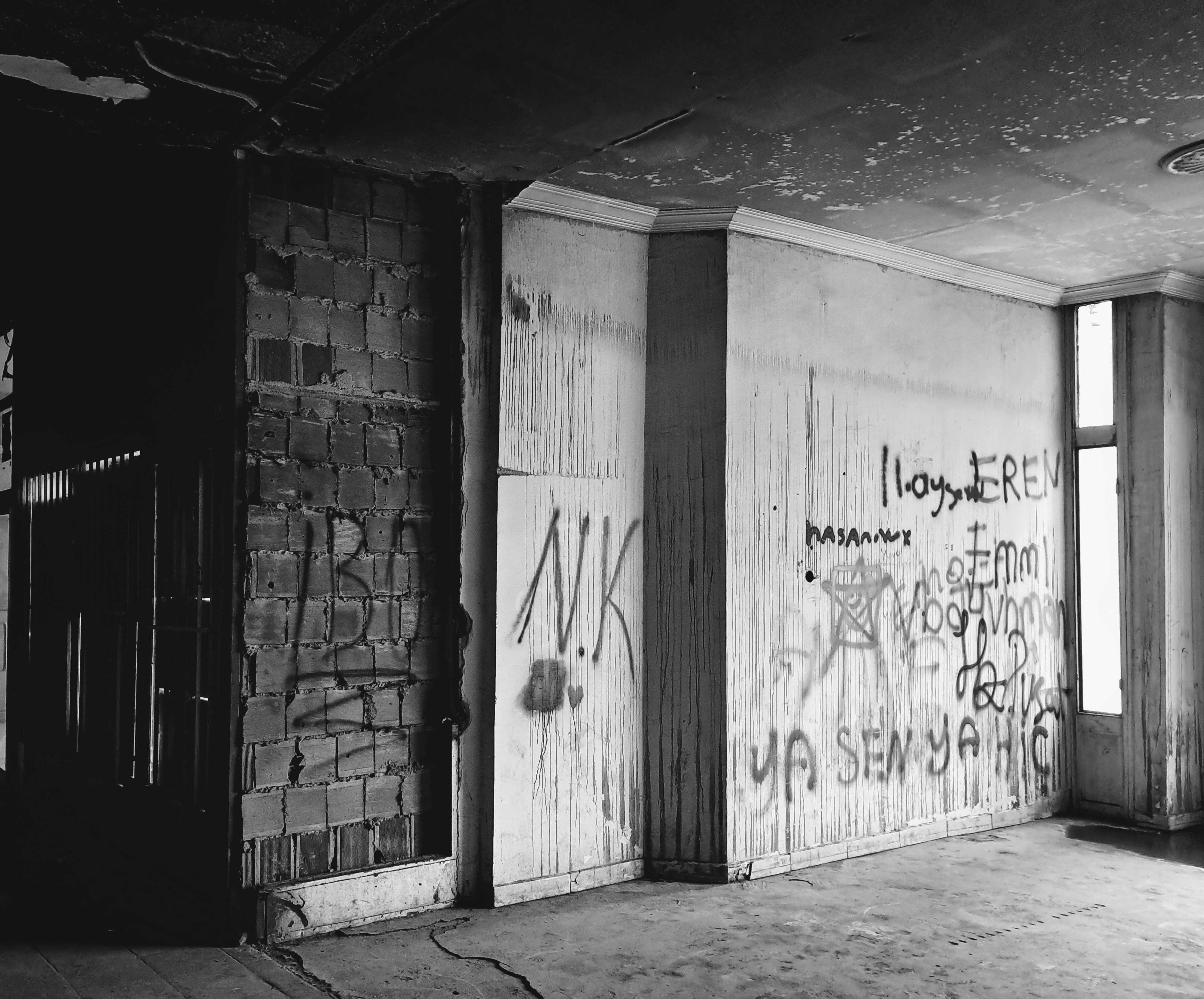 A dilapidated empty hall with a peeling ceiling and a section of exposed brick wall on the left. The white plastered walls on the right are covered in various dark graffiti, with legible text including "N.K", "EREN", "WX", "Unc", and "ITS YOU OR NOTHING". There are many other overlapping scribbles and marks that are difficult to decipher. A narrow window on the right lets in daylight, contrasting with the deeply shadowed opening on the left.