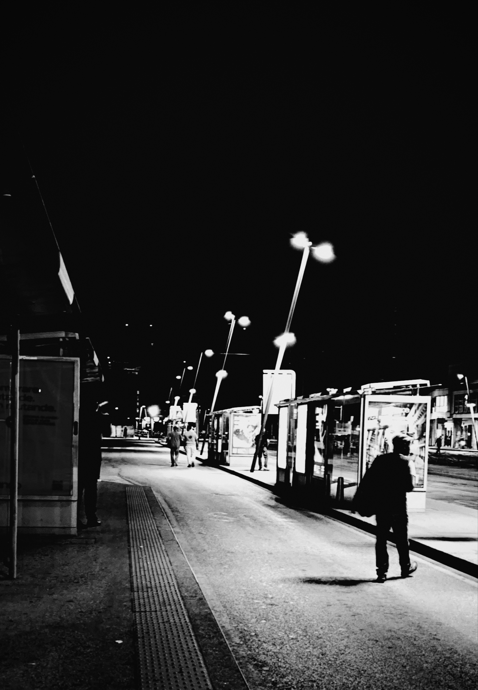 Persone si muovono tra i marciapiedi che servono la fermata di alcuni autobus. La foto è scattata in bianco e nero di notte, sotto alcuni lampioni.